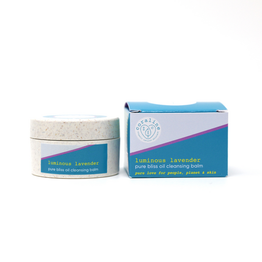 A jar of Coraline skincare's "Luminous Lavender" pure bliss oil cleansing balm is pictured next to its box. The jar has a speckled beige lid and white label with purple and green accents. The box is turquoise and white with similar coloured accents and text.