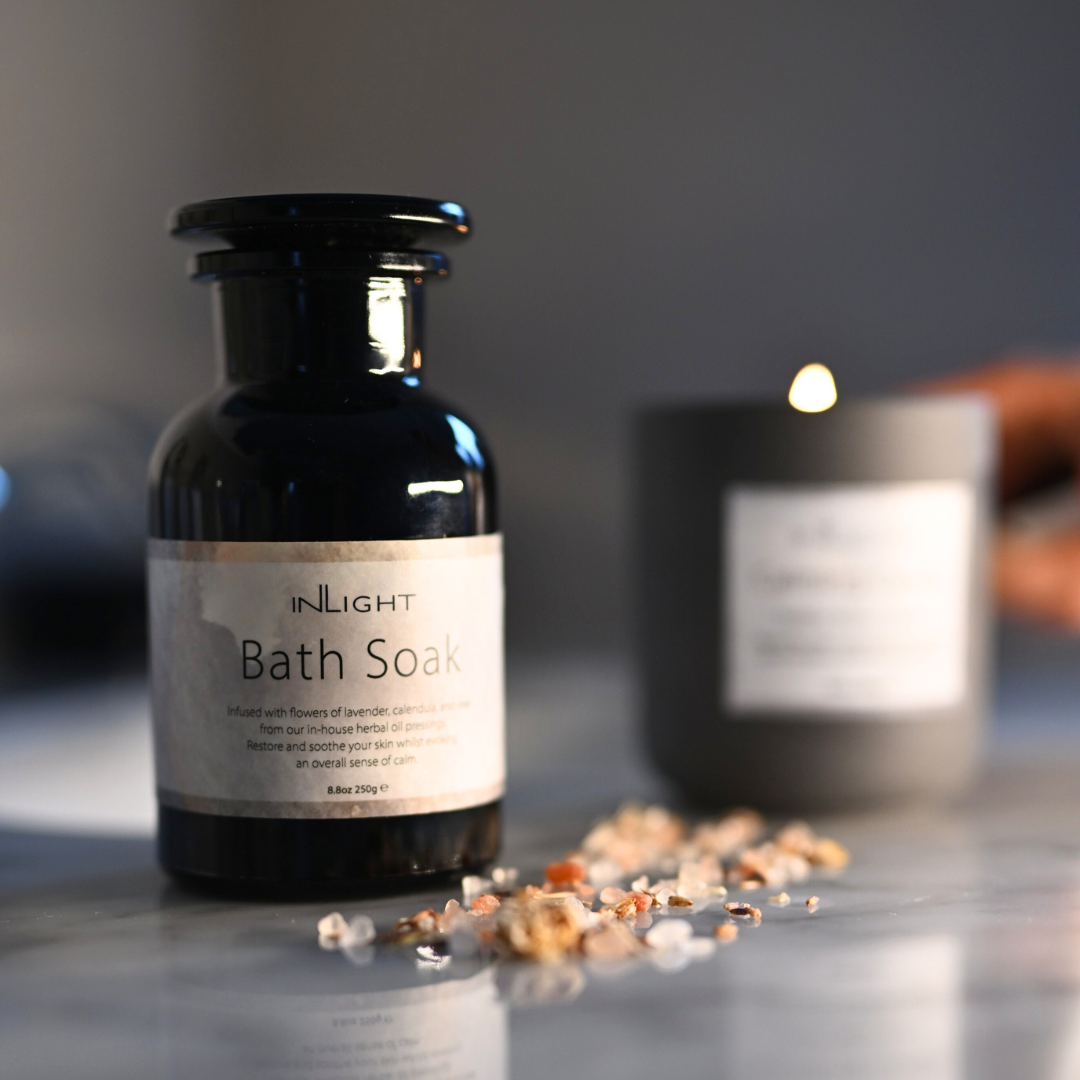 Bottle of 'INLIGHT Bath Soak' with a blurred candle in the background on a reflective surface. Salts and dried florals are scattered in the foreground and you can see a lit candle out of focus in the background