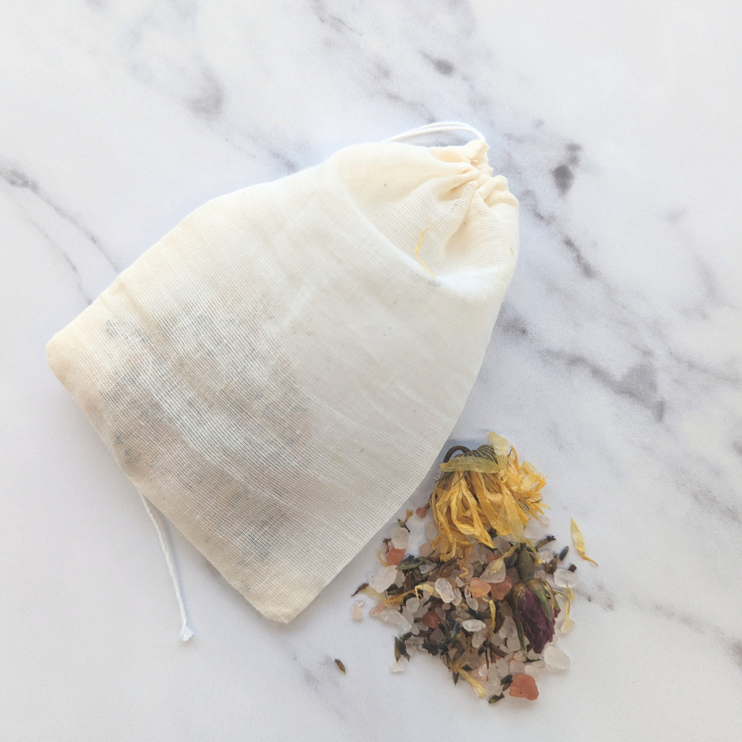 white and grey worktop where an organic cotton muslin drawstring bag is sitting next to a pile of bath salts and dried herbs and flowers