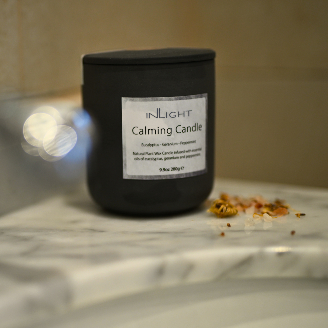 Black calming candle with a label on a marble surface on the edge of a bathroom sink with dried flower buds scattered next to it