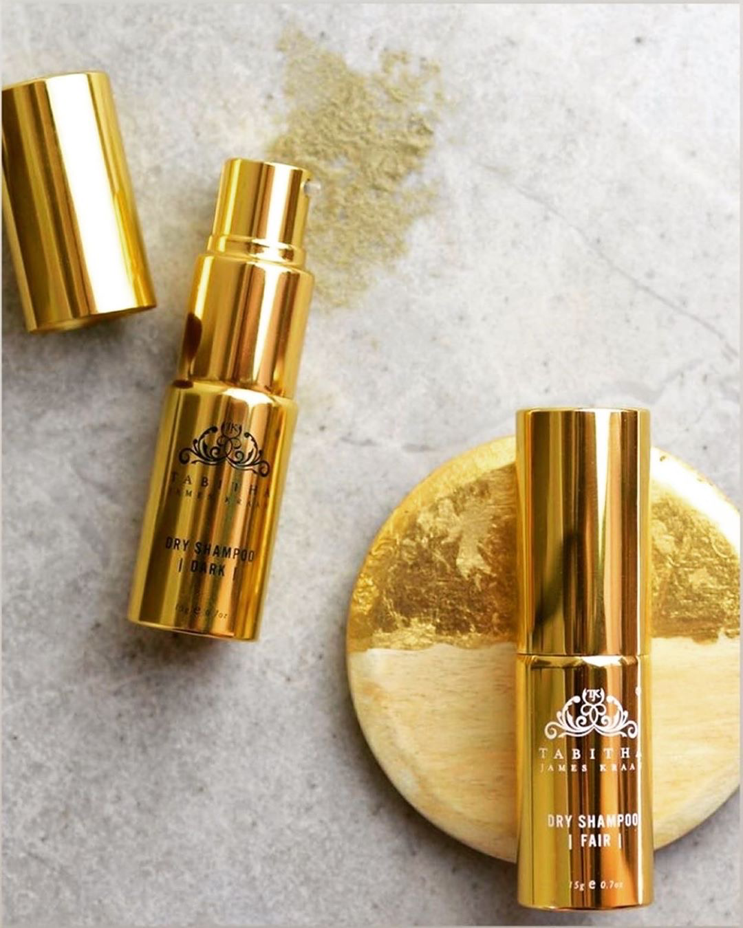 2 travel sized dry shampoos in gold bottles with a pump dispenser. on the left the dry shampoo for brown hair has the cap off and you can see the colour of the powder on the grey tile background. on the right the dry shampoo for blonde or fair hair is laid on a two tone gold coaster