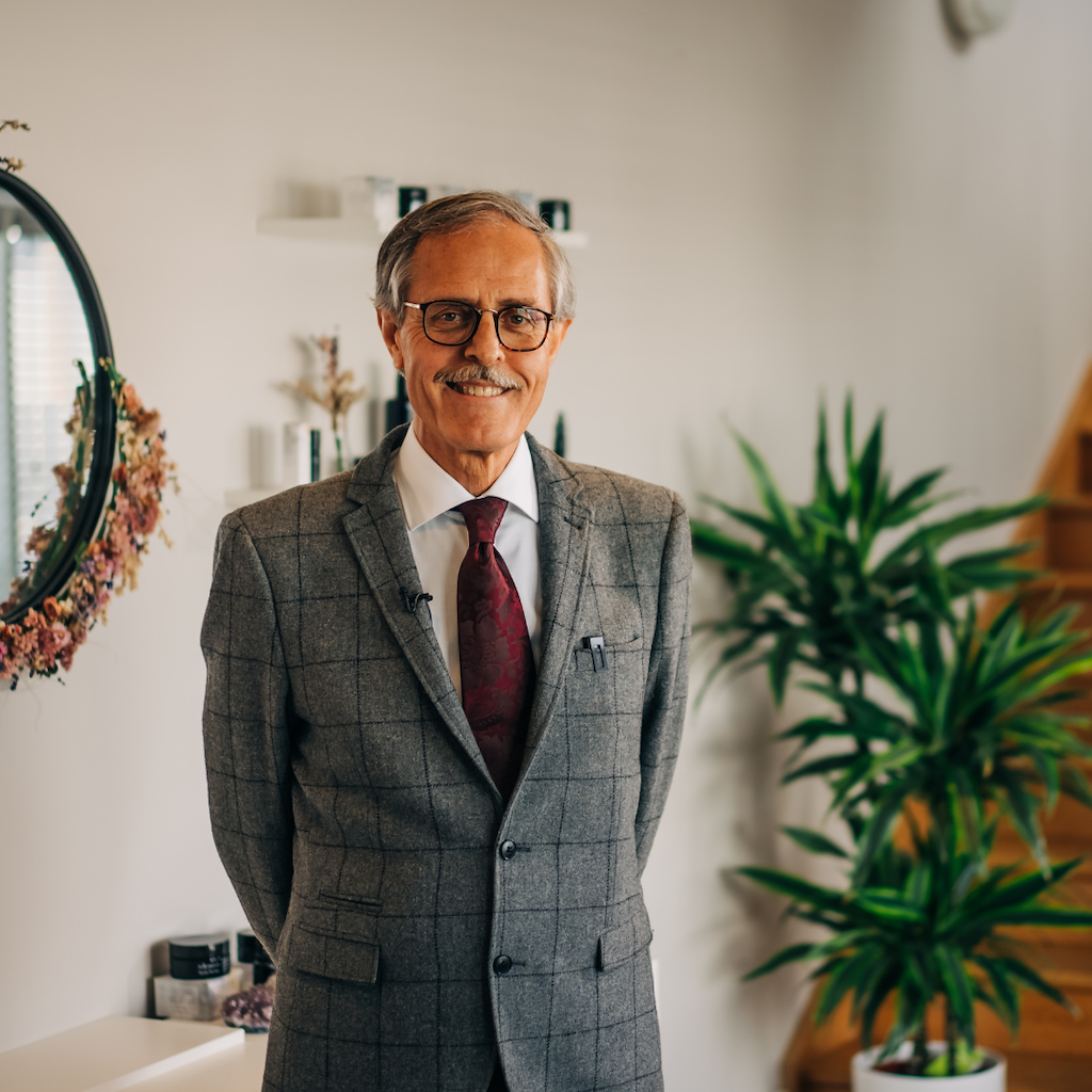 dr spiezia founder of inlight beauty who is standing in a grey checked suit in a hallway with white paint, a black mirror surrounded by pink flowers and a spiky green palm plant and pine coloured stairs in the background