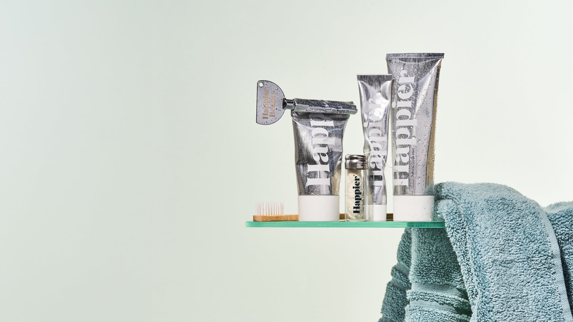trio of natural whitening toothpaste in aluminium tubes on a glass bathroom shelf with floss, toothrush and towel