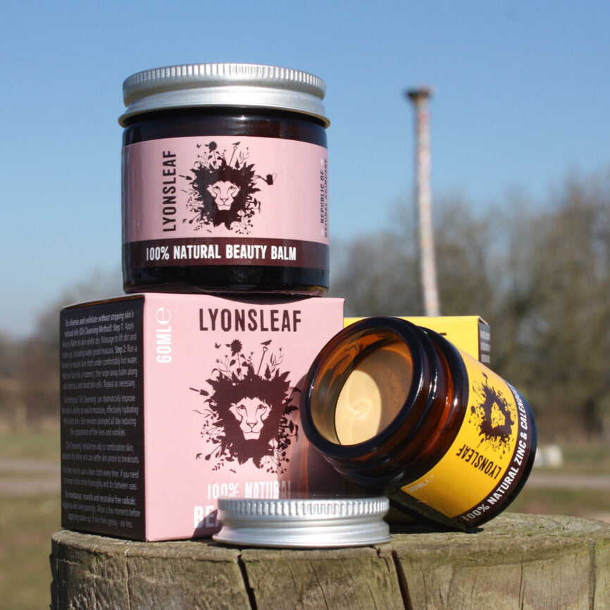 pots of handmade skincare balms sitting on top of colourful pastel cardboard boxes. One jar is open so you can see the yellow creamy texture of the balm inside. The jars and the boxes are sat on a tree stump and in the background there are trees and blue sky