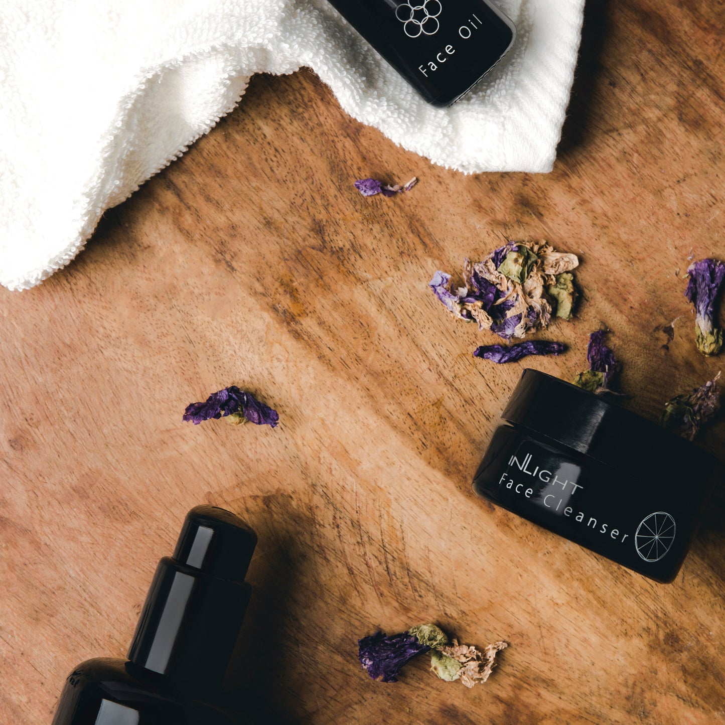 flat lay image of inlight beauty organic skincare products on a wooden table. the black glass cosmetic bottles and jars are surrounded by dried purple flower petals and a white fluffy face cloth