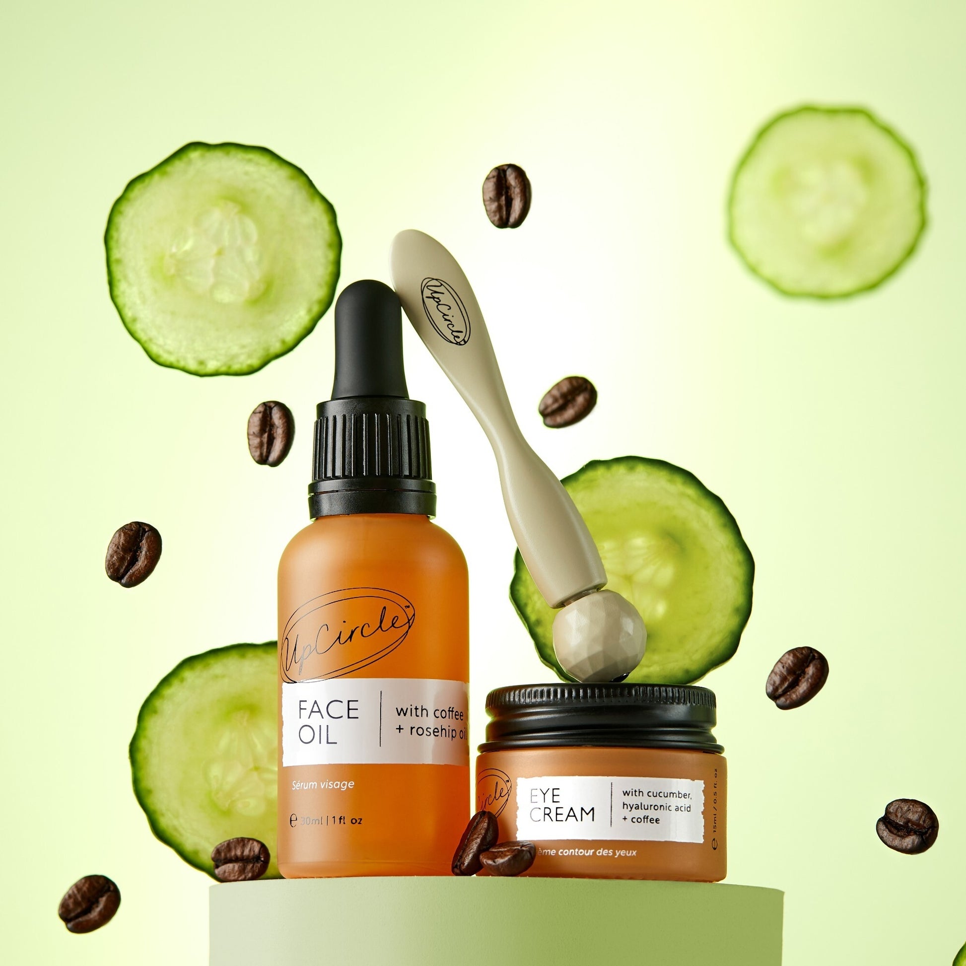 green background with slices of cucumber and coffee beans. in front on a green plinth the products in the caffeinated duo (face oil and eye cream) can be seen as well as the upcircle eye roller