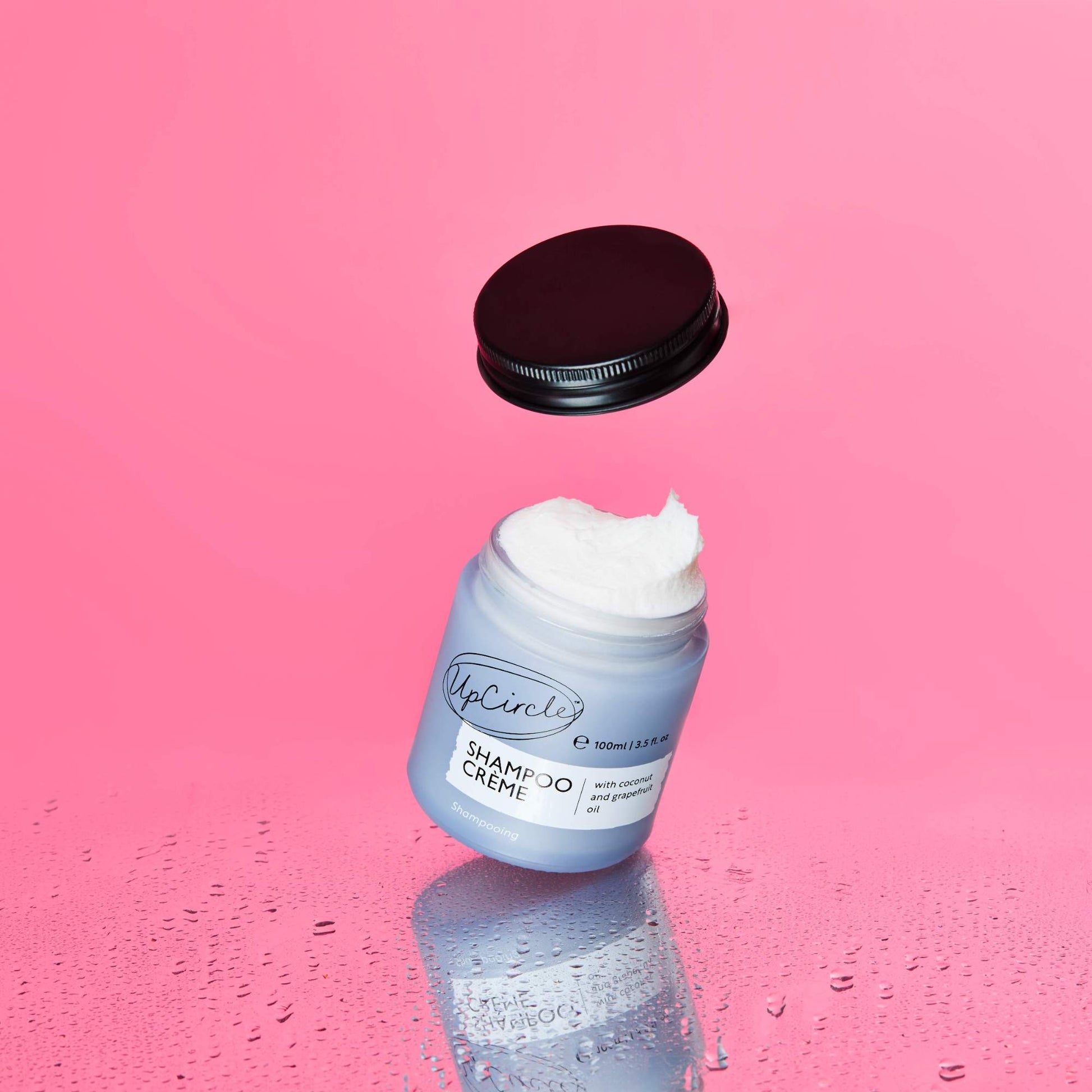 A blue jar of UpCircle Shampoo Crème with the lid floating above it, showcasing a fluffy, white cream texture. The background is a vibrant pink with water droplets on the surface below, creating a fresh and clean aesthetic.