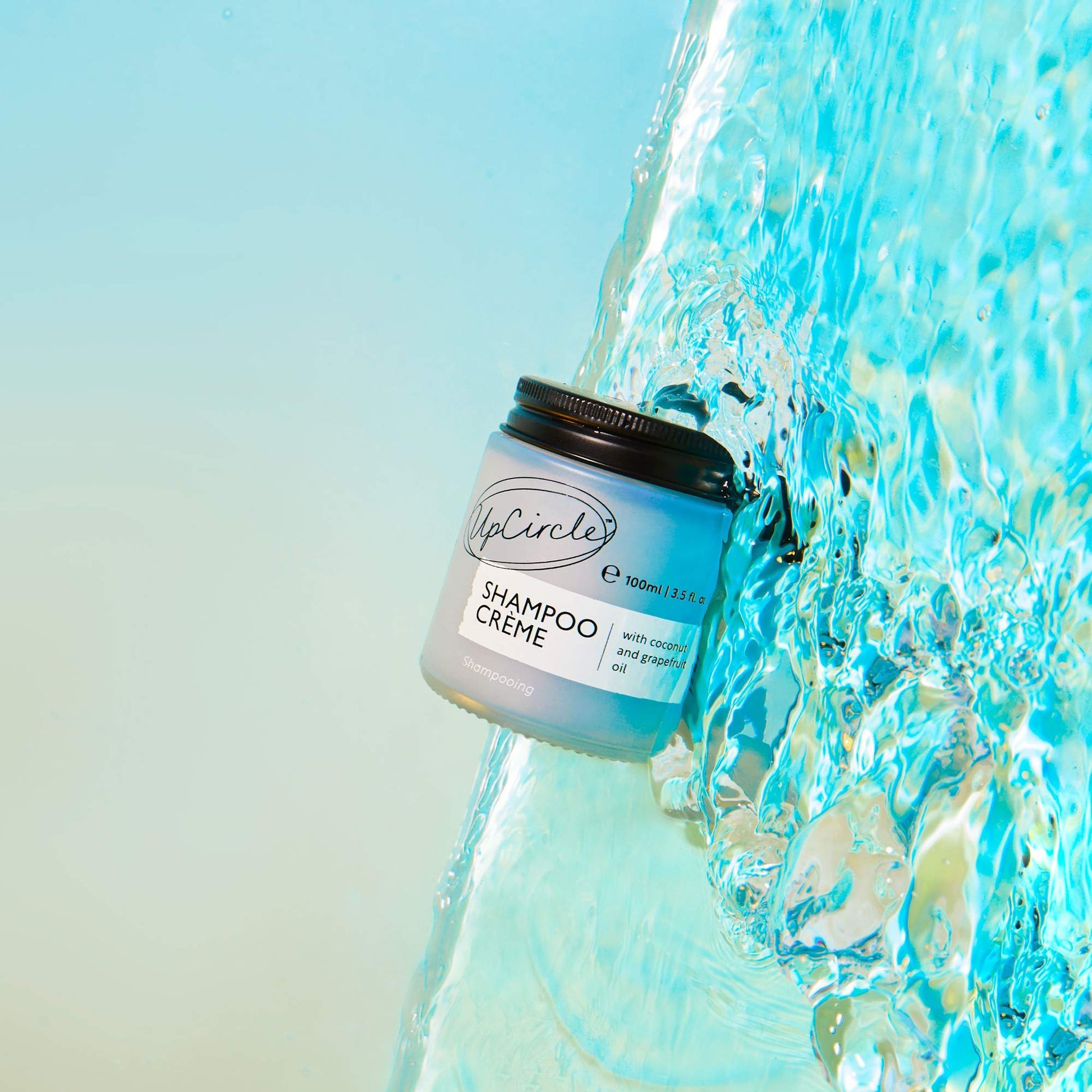 A jar of UpCircle Shampoo Crème with a black lid is pictured against a background of flowing, vibrant blue water. The jar is labeled with the product name and additional information, suggesting it's a revitalizing and environmentally friendly hair care product.