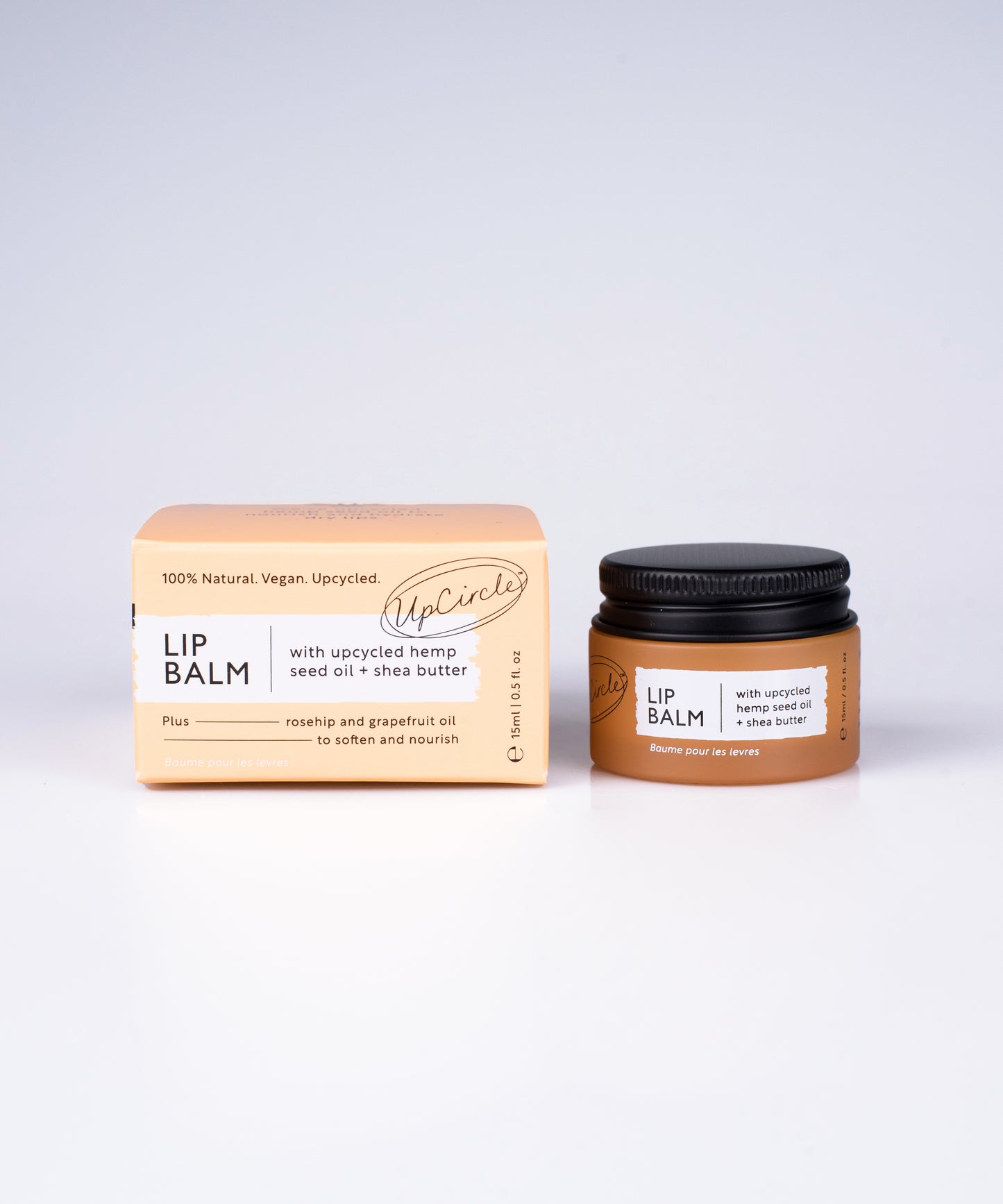 A pot of the upcircle lip balm sits on a plain white background. The orange glass pot is on the right side of the orange cardboard packaging it comes in. The box also lists the brand and product info in black and white text.
