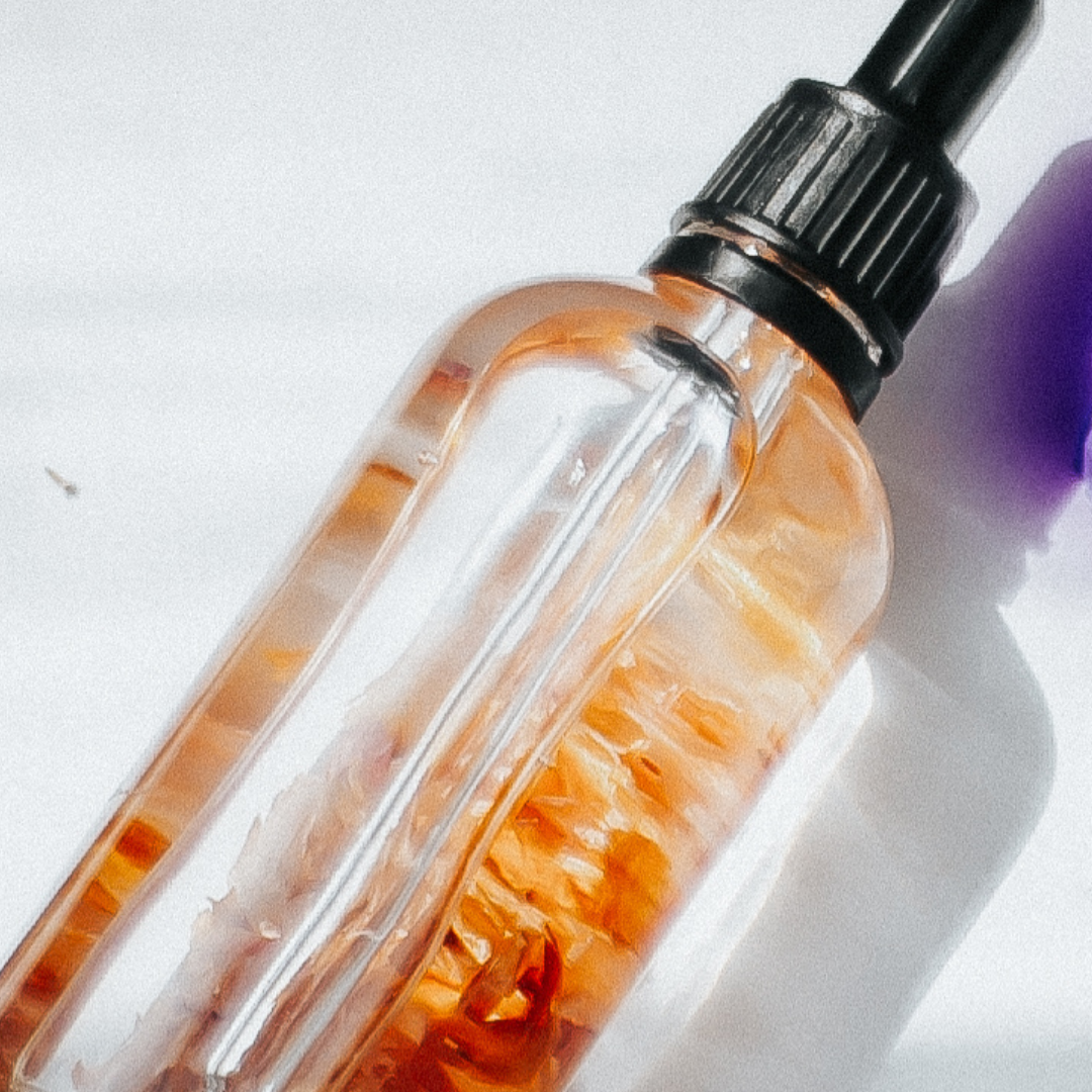 photo of a hand made face oil in a clear glass bottle with black pipette. Inside the oil is lightly coloured and some dried flower petals are suspended within it