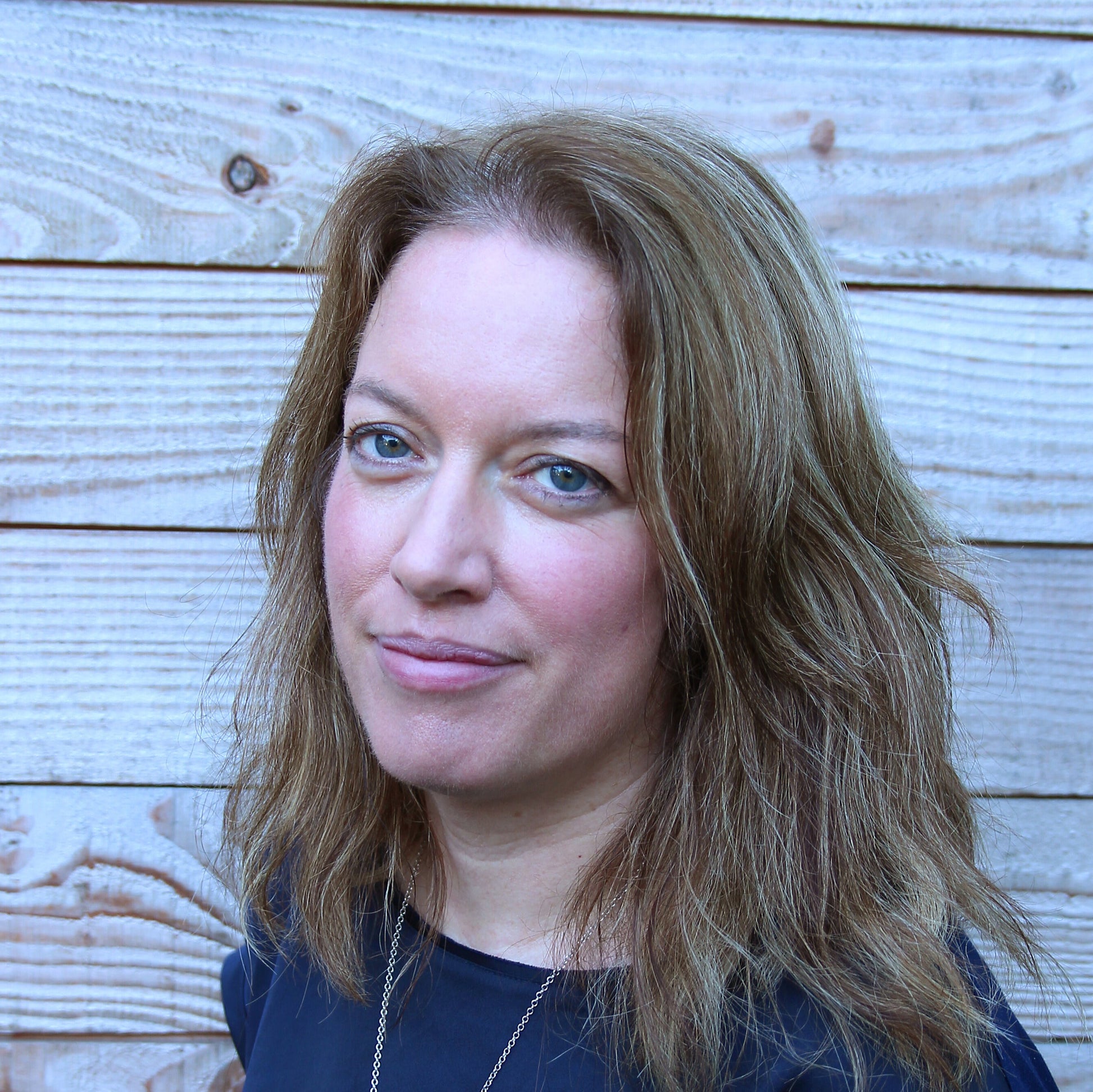 photo of marian who makes silvan skincare vegan face creams from her studio in the welsh borders. Photo of marian standing in front of natural wood panelling with light brown hair and a blue boat neck top with a gold chain