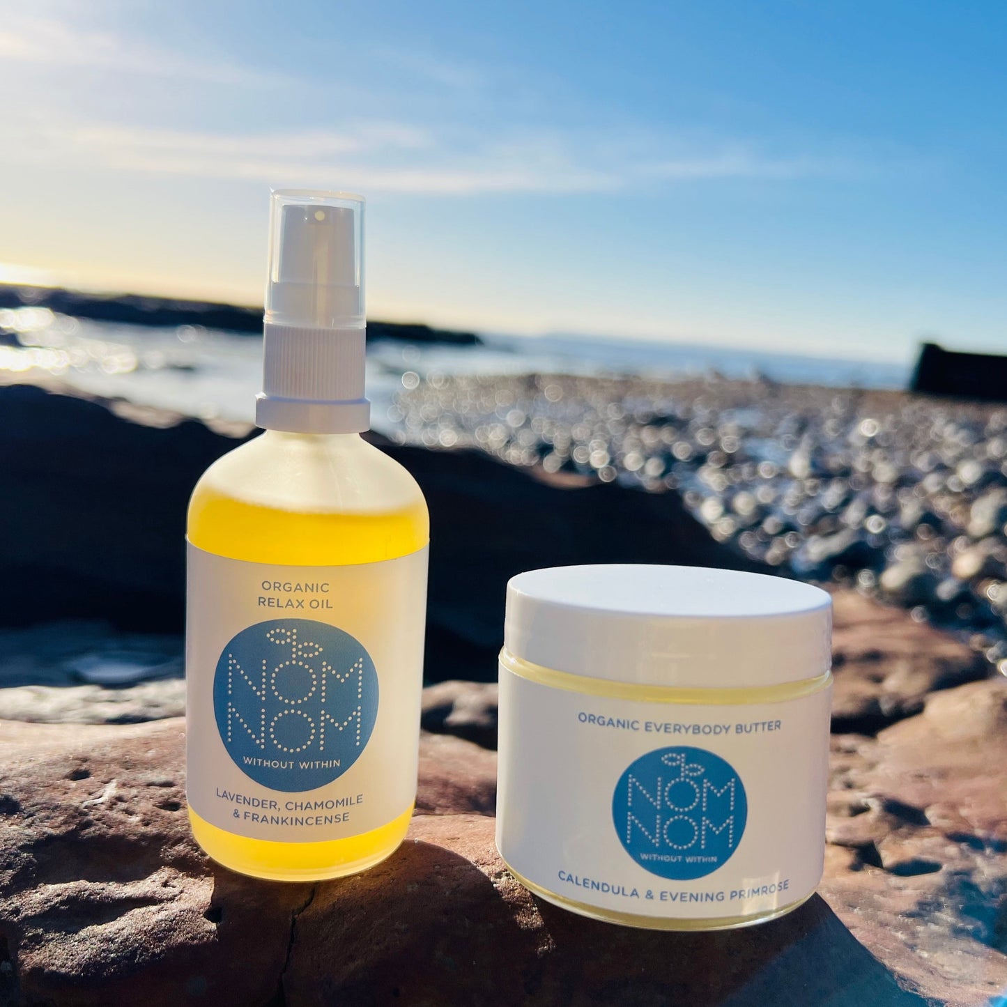 photo of nom nom skincare organic body oil and body butter sitting on the edge of a rock pool. the background is blurred but you can make out a pebble beach and the sea in the background as the products sit in the evening sun