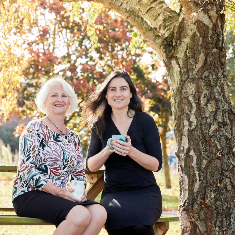 Image of Odylique founders, Margaret Weeds and her daughter Abi, sitting outside on a bench next to tree. 