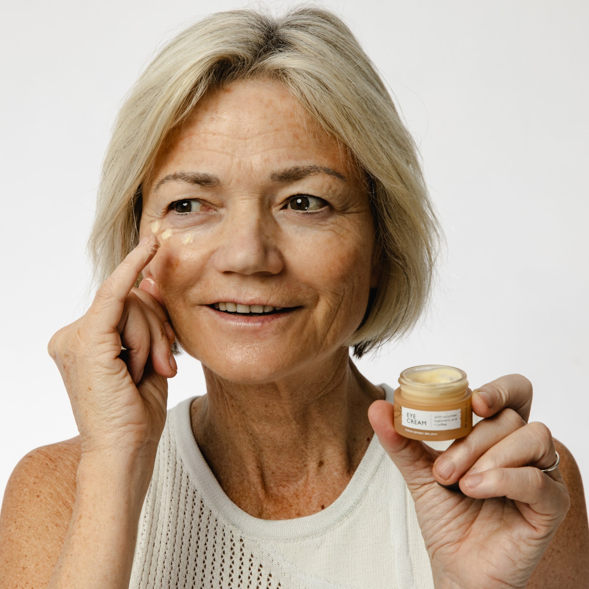 white woman with blonde/white bob applying 3 dots of upcircle eye cream around her eye and holding the jar up to the camera
