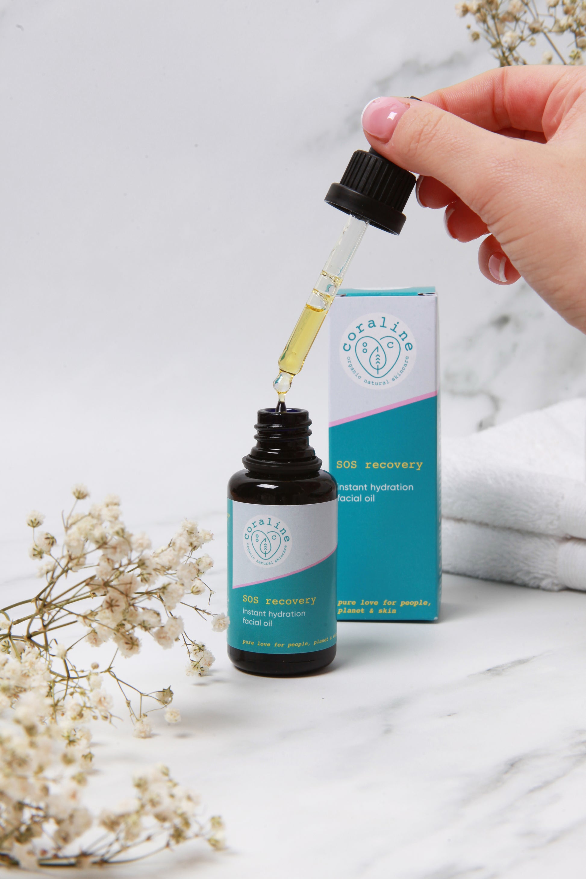 A hand holding a dropper above a small, dark glass bottle of facial oil with a light blue label and a matching box in the background. The dropper has a drop of the oil at its tip. The foreground includes white flowers and the background a folded white towel.