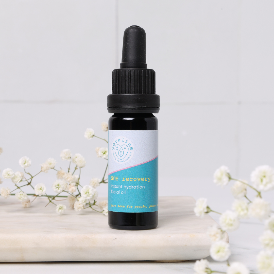A small dark glass bottle of SOS recovery oil with a light blue label. The bottle sits on a marble surface with baby's breath flowers around it