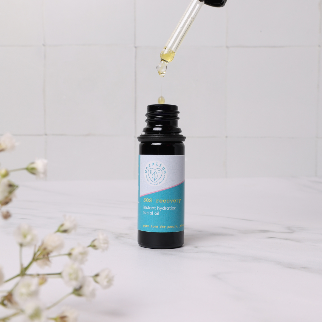 A small dark glass bottle of SOS recovery oil with a light blue label. The dropper has a drop of the oil at its tip. The foreground includes white flowers.