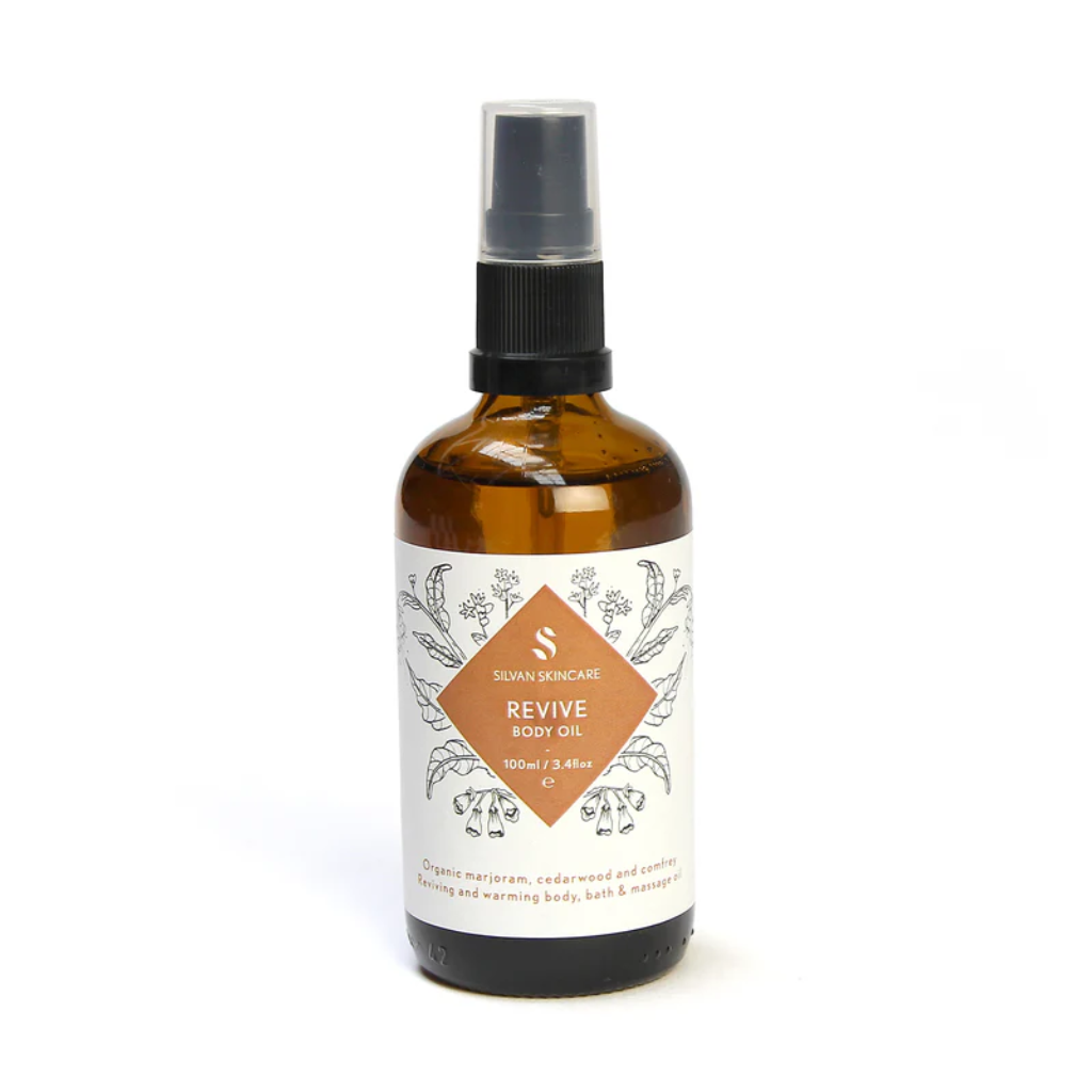 Silvan Skincare revive body oil in an amber glass bottle with a white label. In the centre is a dark orange diamond listing the product and brand. The label also has light black botanical illustrations.