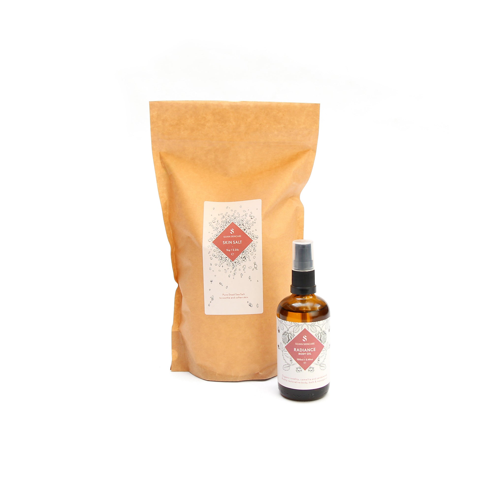 A brown bag of the skin salt and the radiance Body oil placed in front of it, slightly to the left. Both items are have a white label and a pink diamond in the middle, listing the product and brand name.