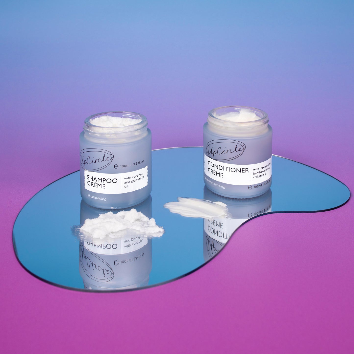 upcircle's hair care duo lifestyle image. the shampoo creme and conditioner creme are sitting on a teardrop shaped mirror and the background is a blue pink ombre colour. the hair care products are shown in their light blue/purple glass jars without lids so you can see the texture of the products. the creams are also spread on the mirror