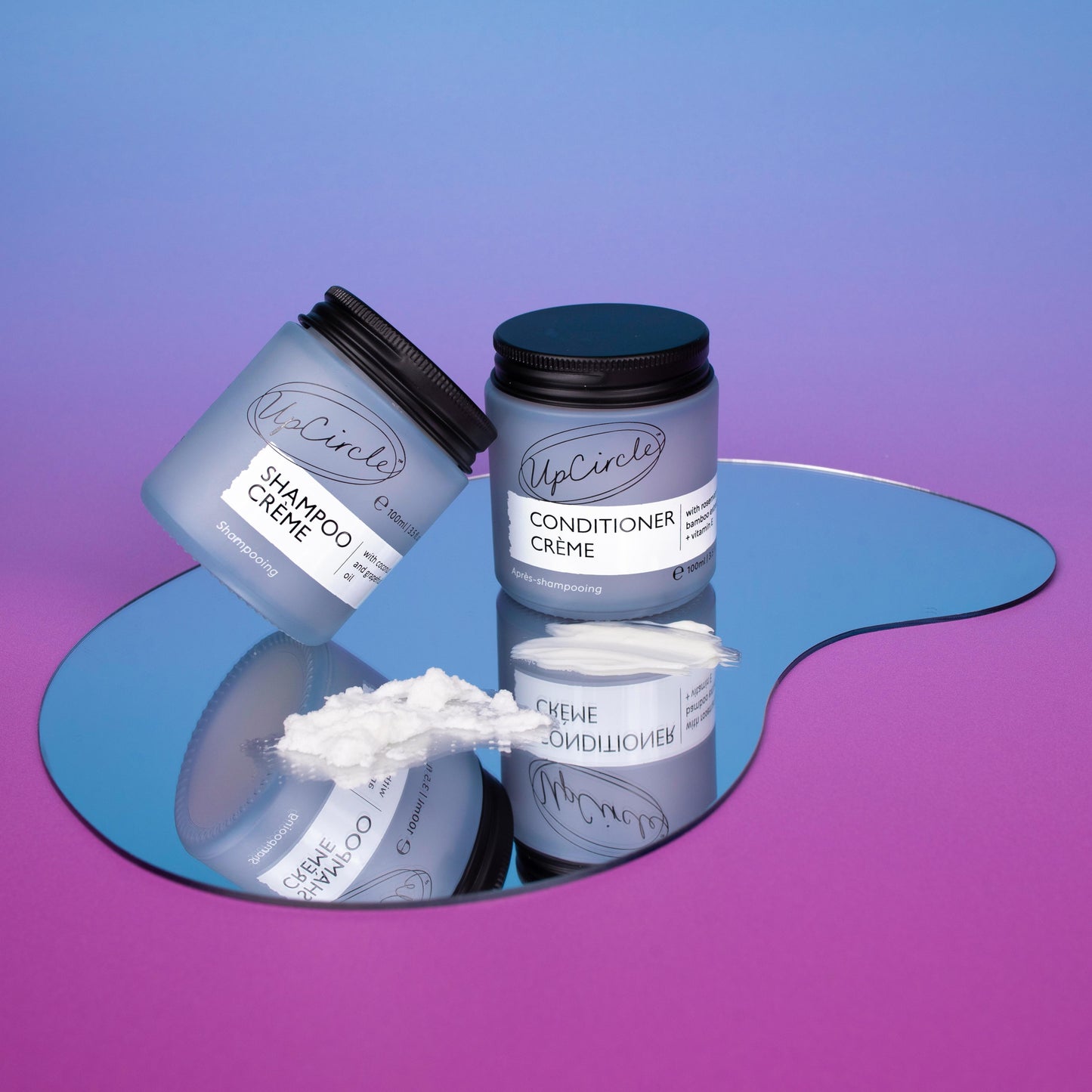 upcircle's hair care duo lifestyle image. the shampoo creme and conditioner creme are sitting on a teardrop shaped mirror and the background is a blue pink ombre colour. the hair care products are shown in their light blue/purple glass jars and the creams are also spread on the mirror