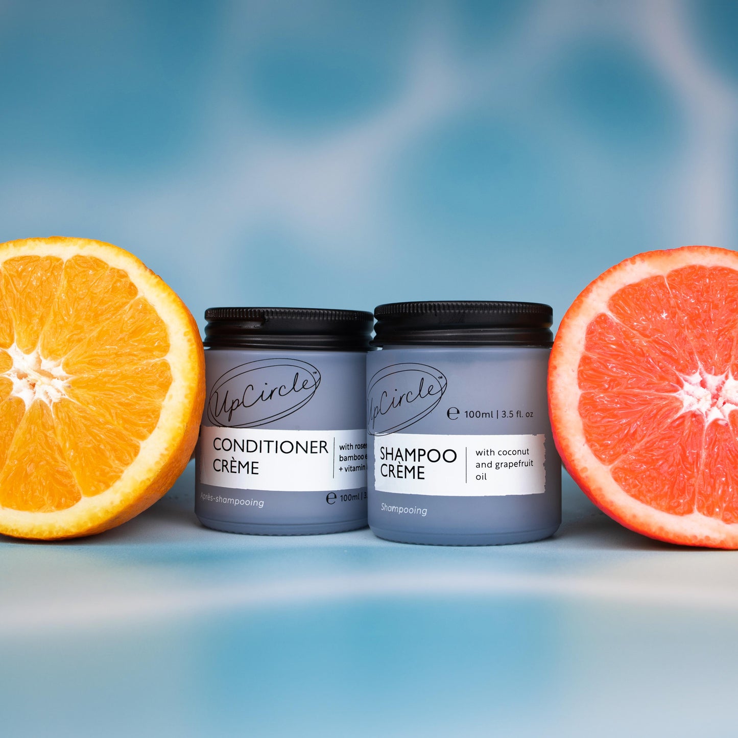 lifestyle photo of upcircle's shampoo creme and conditioner creme on a blue iridescent background. both products are in light blue glass jars with black aluminium lids. there are slices of orange and grapefuits in the background