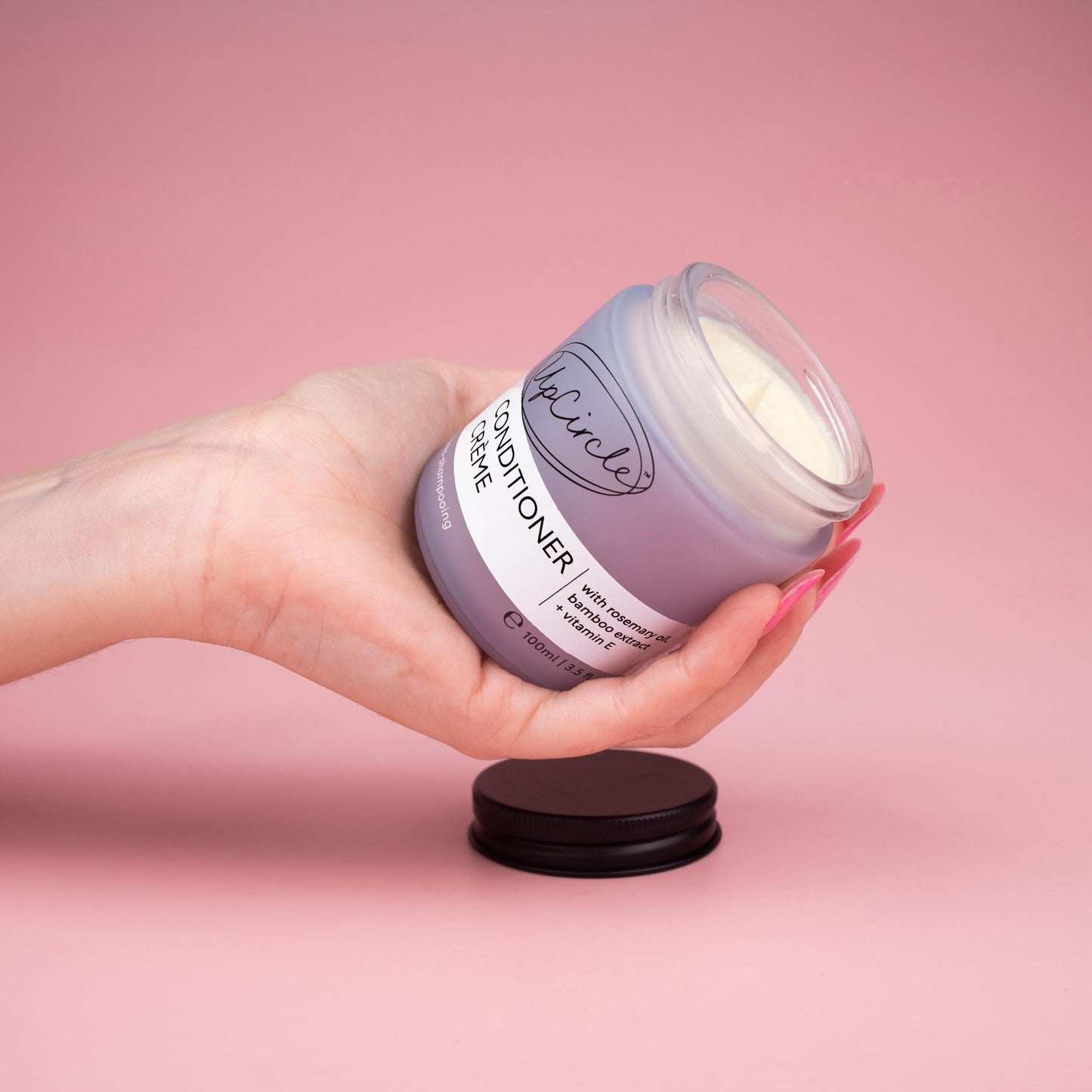lifestyle photo of upcircle's conditioner cream on a pink background. the conditioner is in a blue/lilac frosted glass jar with black aluminium lid. The jar is open and you can see the creamy texture of the conditioner inside. It is being held by a woman's hand with multi tonal pink finger nails