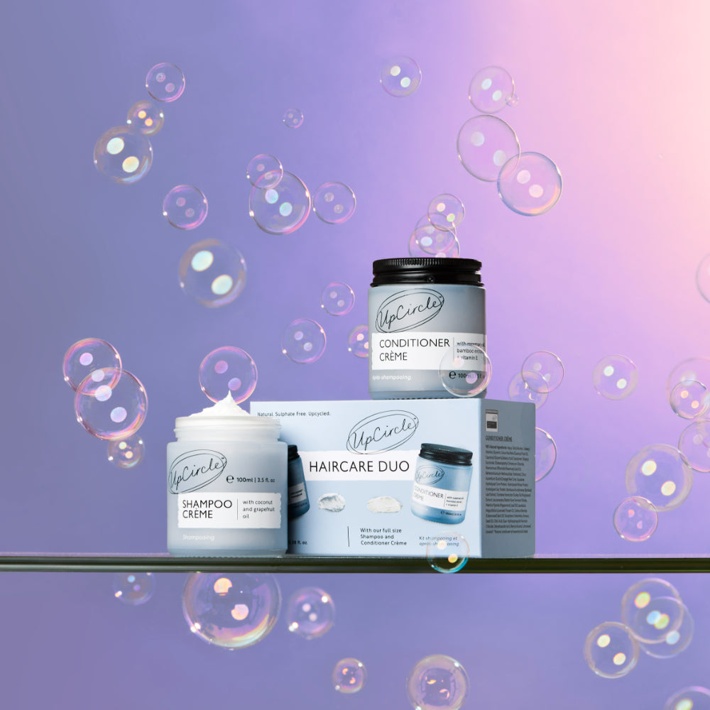 lifestyle image of upcircle's hair care duo box on a purple and pink ombre background. the conditioner is sitting on top of the cardboard box and the shampoo creme is in the blue glass jar with the lid off and the white cream is whipped up and peeking over the top of the jar