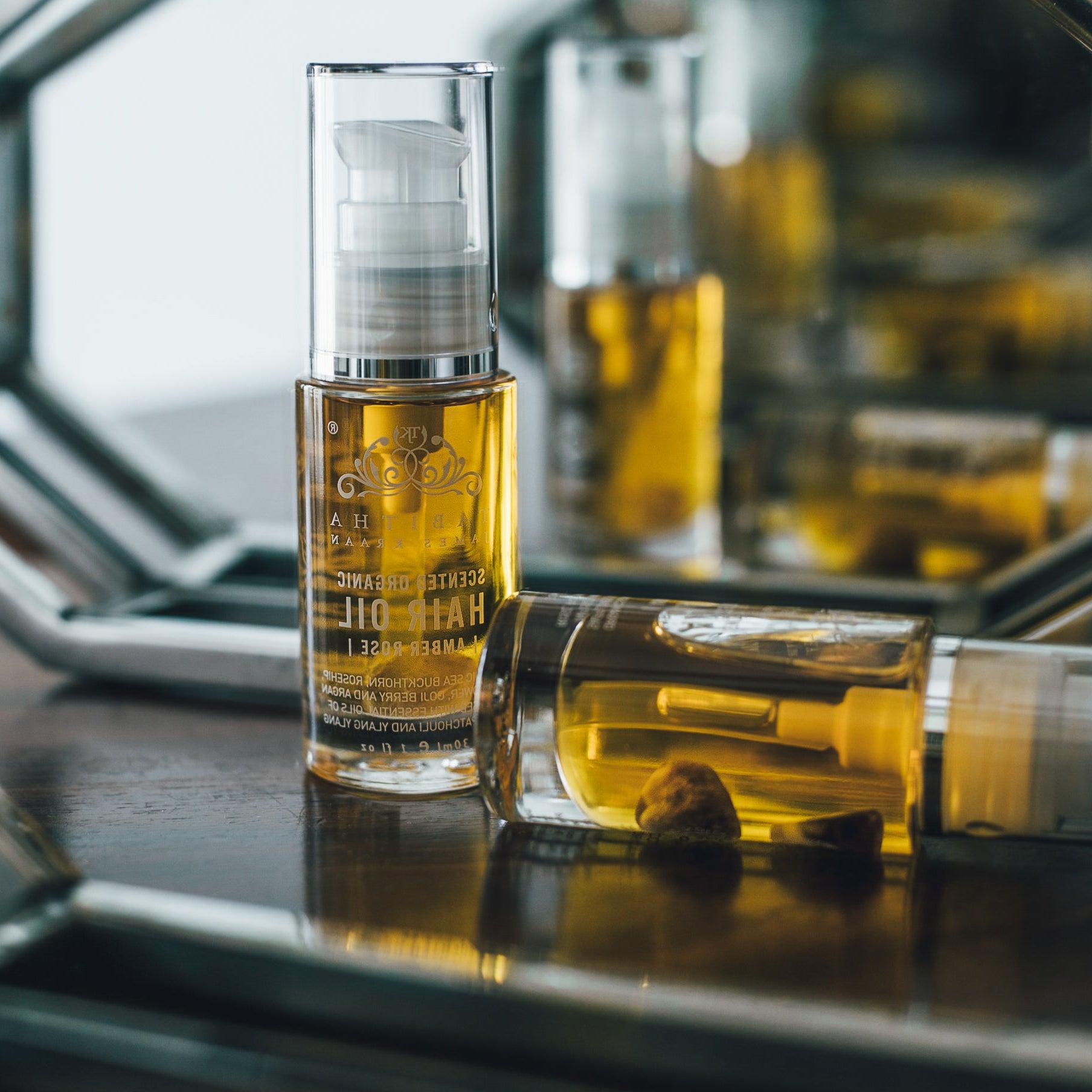 two bottles of hair oil on a dressing table with mirror in the background. the oil is a golden colour and there is a amethyst crystal inside the bottle