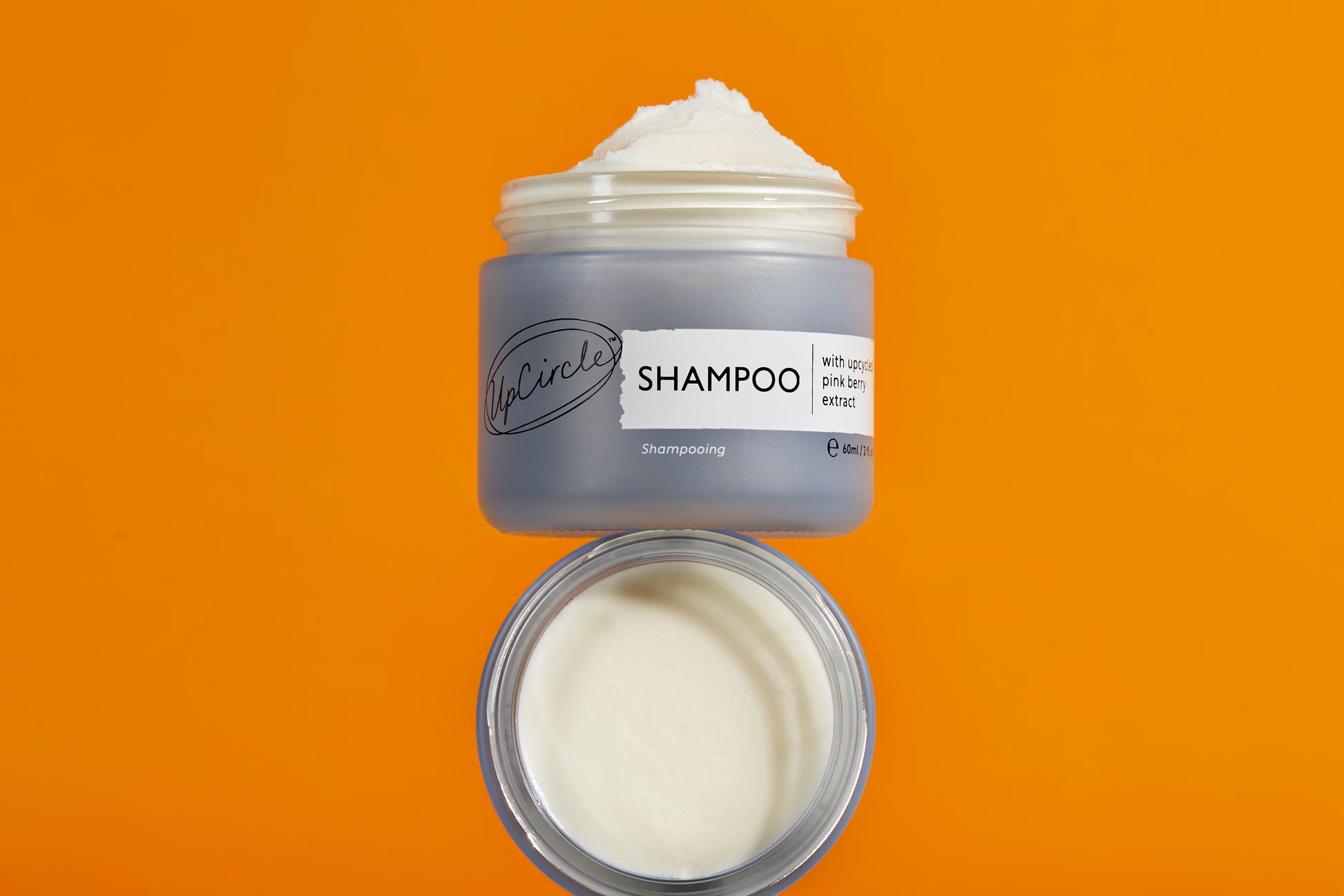 A jar of upcircle shampoo with an open lid shown against an orange background. The jar contains the white, creamy product and sits a top another jar that is balanced on its side.