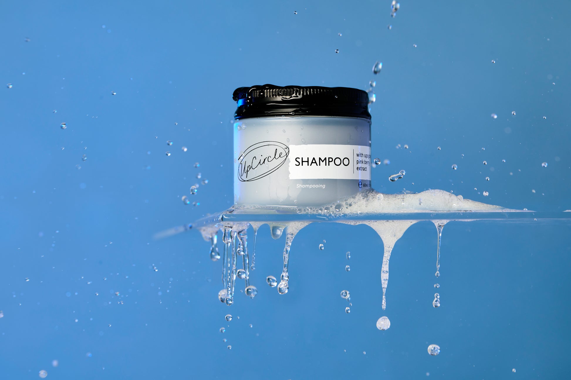 A jar of shampoo labeled "Shampoo" with black lid and white body, by the brand "Upcircle," sits on a wet, partially submerged surface against a gradient blue background. Water droplets and a bit of foam are visible, adding a fresh and clean vibe to the image.