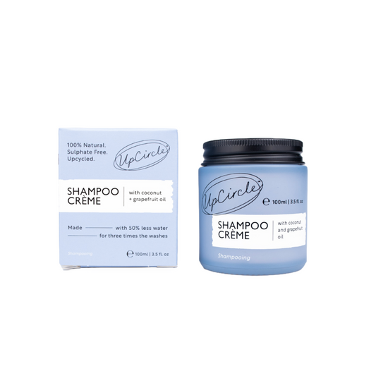 A jar of UpCircle Shampoo Creme with coconut and grapefruit oil is placed next to its box. The jar is light blue with a black lid, and the box highlights that the product is 100% natural, sulfate-free, and upcycled. The jar holds 100ml or 3.5fl oz.