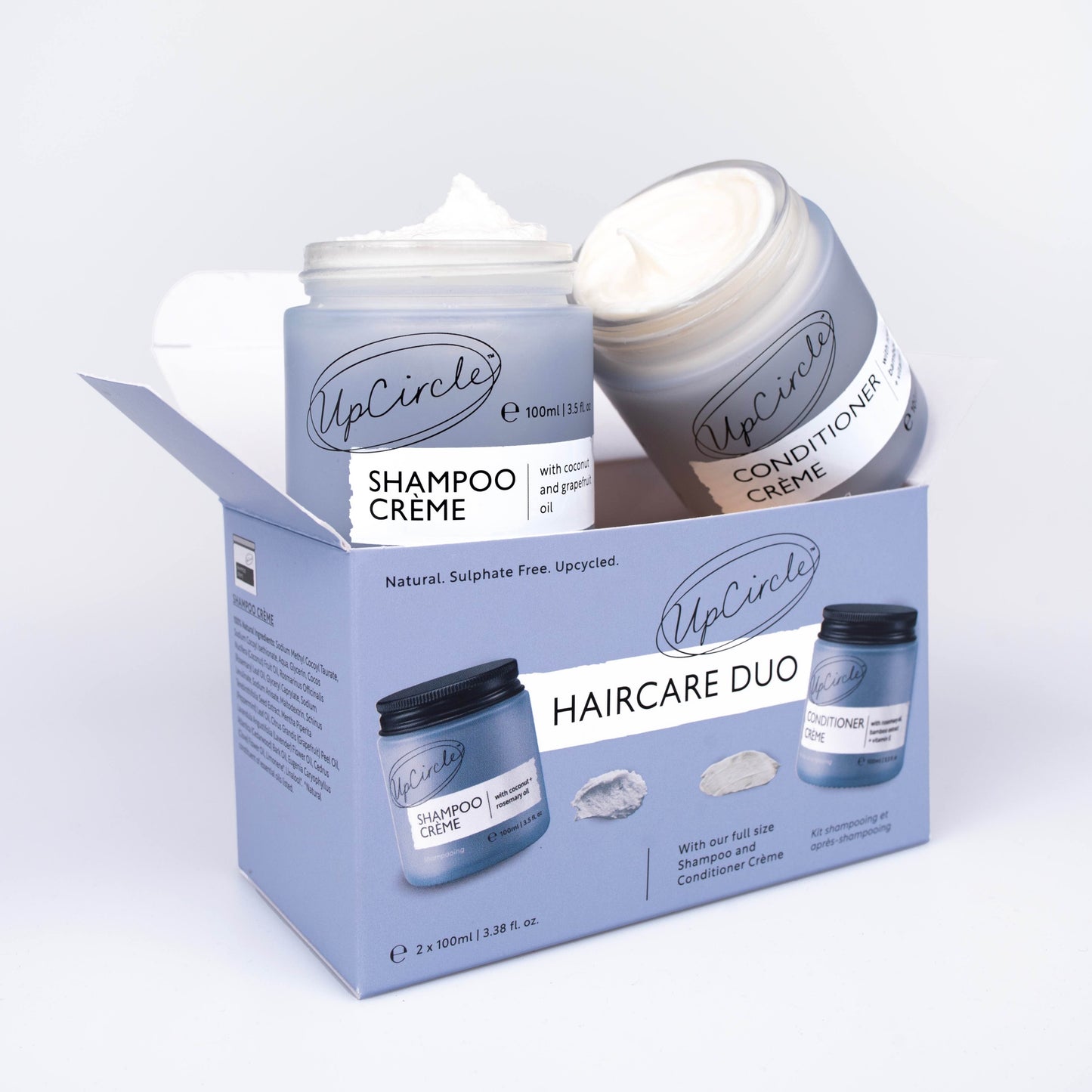 upcircle beauty hair care shampoo and conditioner duo on a white background. The shampoo and conditioner are sitting on top of the outer box. The jars are open and you can see the creamy texture of the hair products inside the jar.The box is a blue/lilac colour to match the frosted glass jars which have black aluminium lids