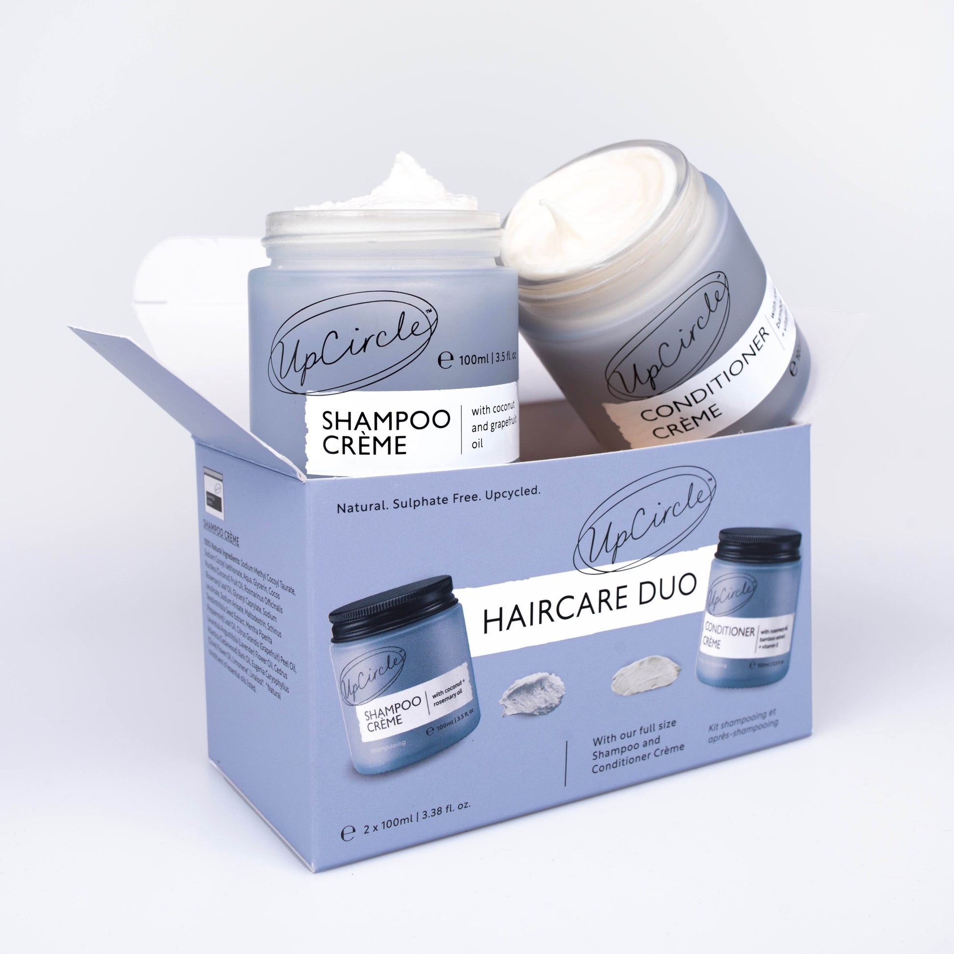 upcircle beauty hair care shampoo and conditioner duo on a white background. The shampoo and conditioner are sitting on top of the outer box. The jars are open and you can see the creamy texture of the hair products inside the jar.The box is a blue/lilac colour to match the frosted glass jars which have black aluminium lids