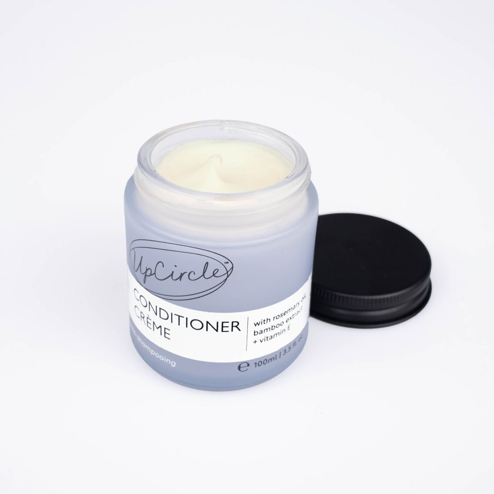 photo of upcircle beauty's conditioner creme on a white background. the hair conditioner is in a blue/purple glass jar with black aluminium lid which is sitting next to the jar and you can see the yellow/cream colour of the conditioner inside. The white label reads 'upcircle conditioner creme with rosemary, bamboo extract + vitamin e' 100ml