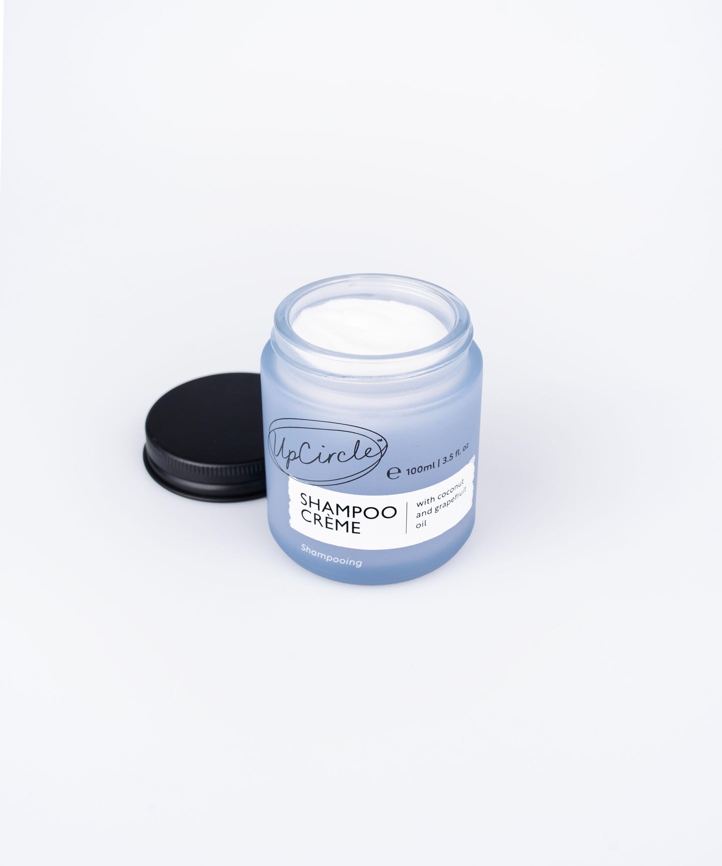 A blue jar with a black lid next to it, containing UpCircle Shampoo Creme. The jar label reads "Shampoo Creme 100ml/3.5fl oz, with mandarin and chamomile." The lid is off, revealing the white shampoo creme inside. The background is white.
