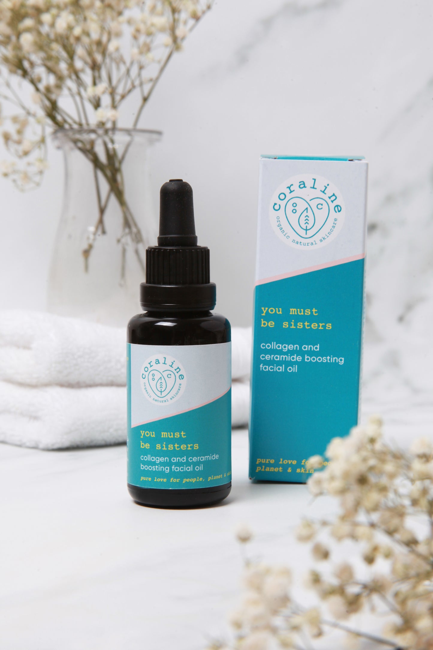A bottle of "You Must Be Sisters" facial oil stands in front of its packaging on a marble surface. Behind them, white towels and delicate baby's breath flowers in a vase. The flowers are blurred out in the foreground too.