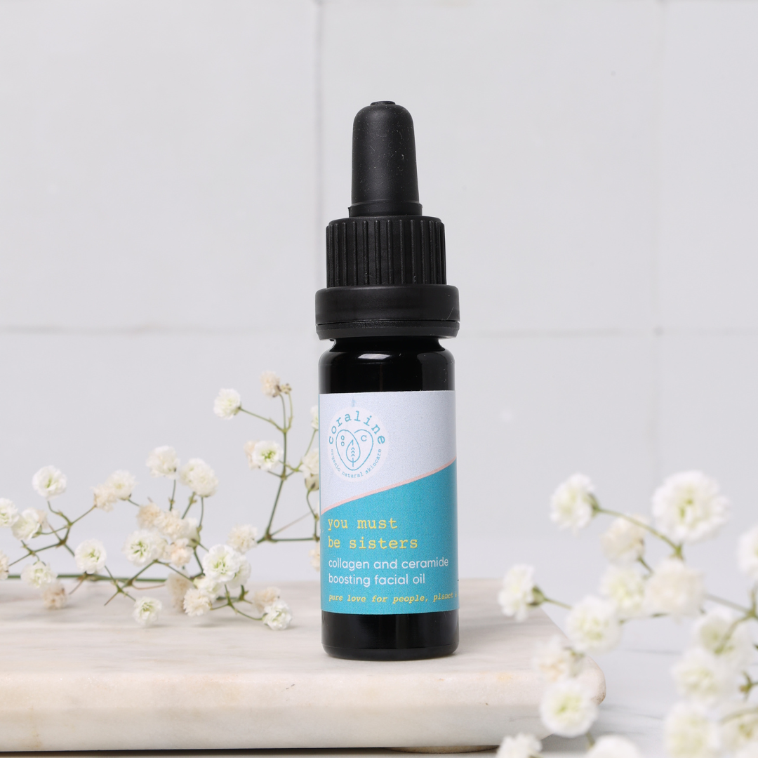 A bottle of "you must be sisters" collagen and ceramide-boosting facial oil. The bottle has a blue and white label. In the foreground, there's a small sprig of white flowers on a marble surface.