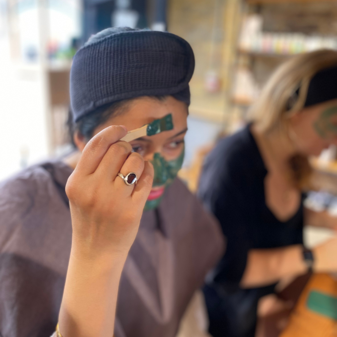 A person is holding a small brush and using it to apply a green face mask to their forehead. They are wearing a black headband and a purple gown. Another person in the background is also wearing the facial mask and a black outfit.