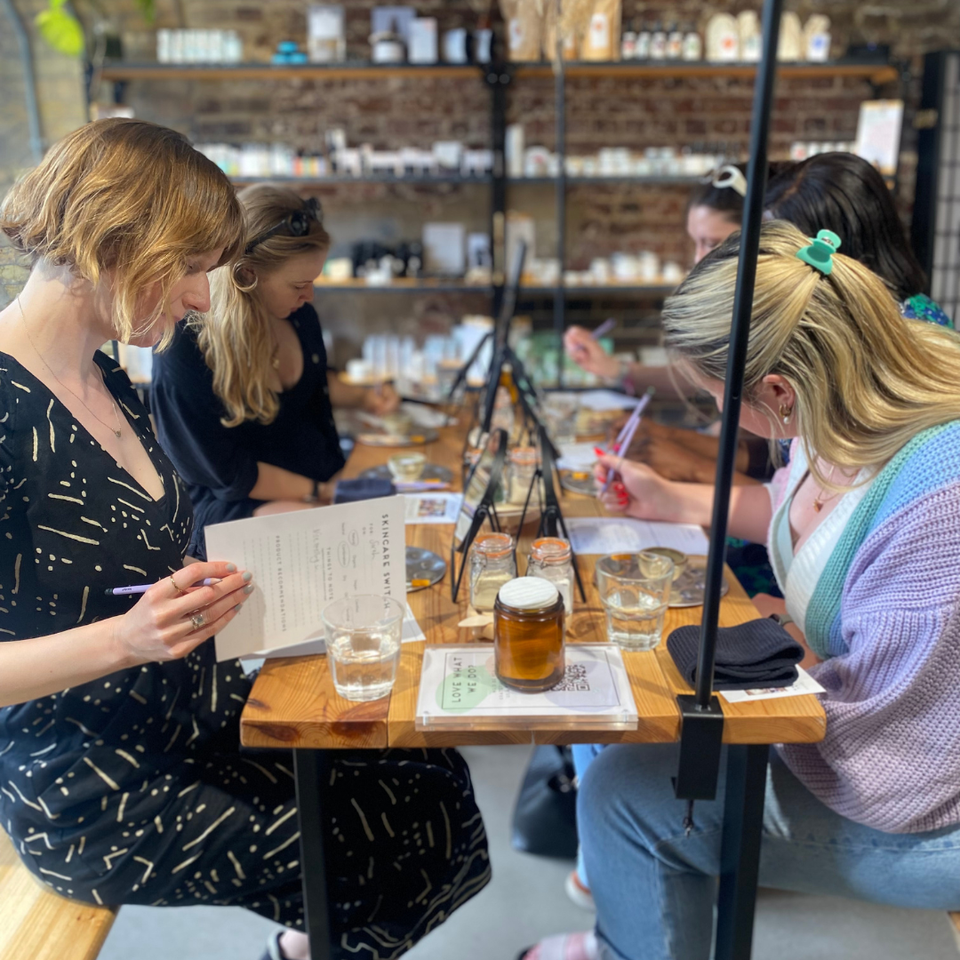 A group of women sit at a wooden table on the cozy Blomma shop floor, engaged in a workshop. They are focused on learning, with various supplies and jars on the table. Shelves of the shops clean beauty products line the walls in the background.