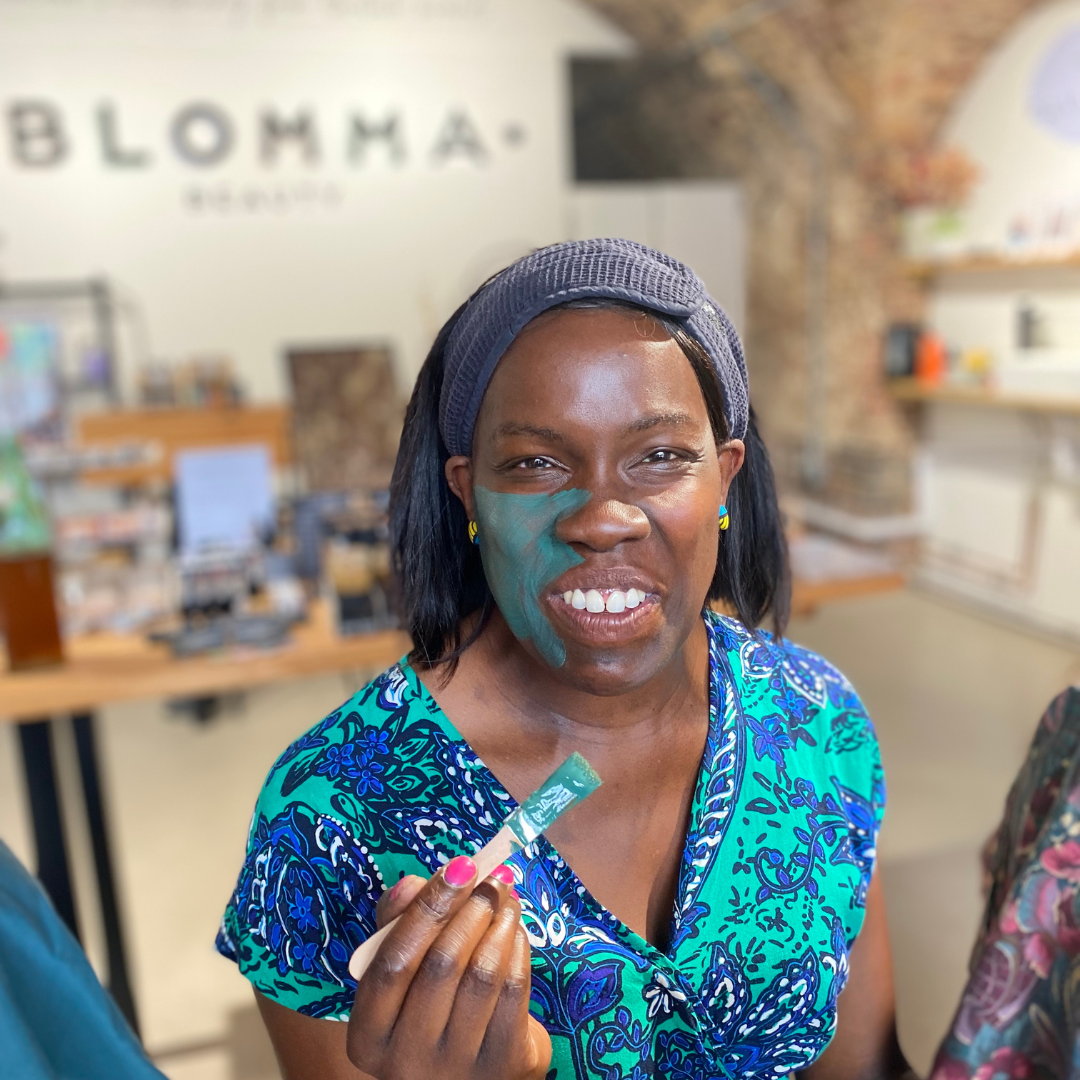 A woman wearing a colourful floral dress and a gray headband holds a small face brush. She has green face mask on her cheek and is smiling.