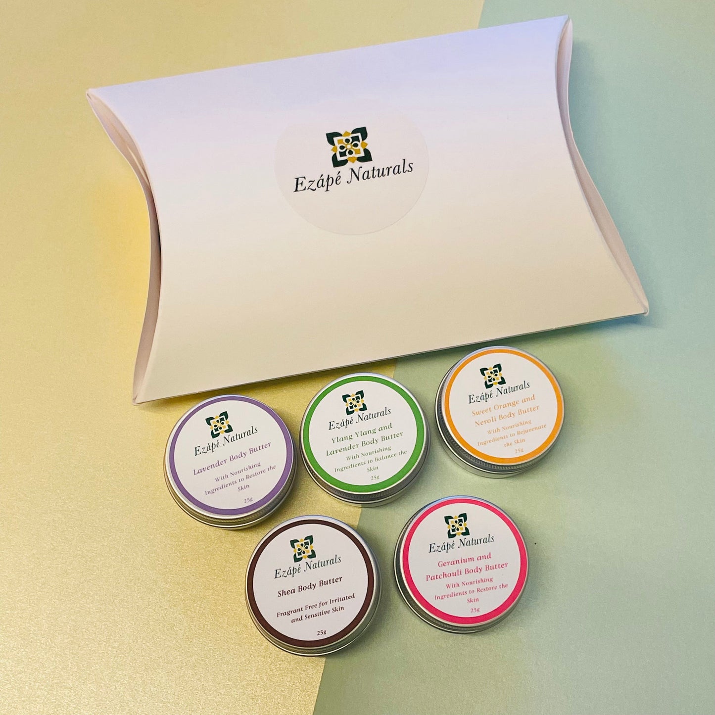 a white pillow pack with the ezape naturals logo on it and underneath there are 5 tins of hand made body butter each with a colourful label to denote the different scents: lavender (purple), lavender and ylang ylang (green), sweet orange and neroli (orange), shea butter (brown), geranium and patchouli (pink)