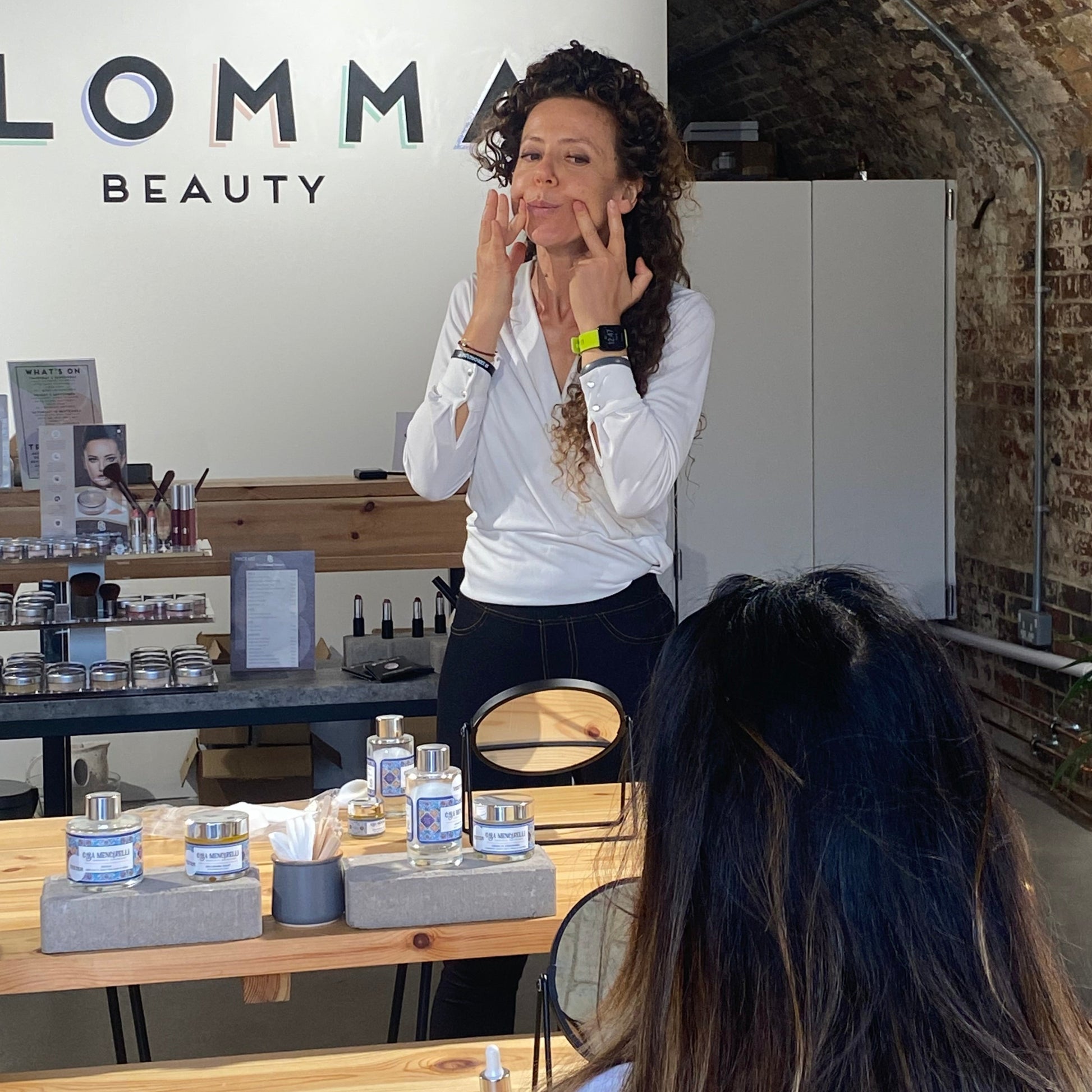 lucia mencarelli is teaching a face massage class. She can be seen using her fingers to sculpt her face. the back of a student's head can be seen in the foreground and they are surrounded by organic skincare products