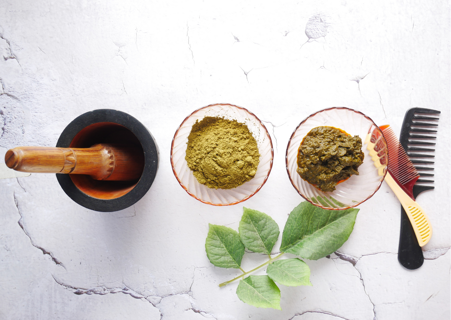 a flat lay lifestyle image of it's pure organic herbal hair dyes. from left to right you can see a wooden and stone pestle and mortar, 2 glass bowls with a pink tint, one showing the dry powder which is an earthy green colour and the other showing the powder mixed with water to form a darker sludgy paste. there is one wooden and one plastic comb to the far right and a green leaf frond at the front of the row