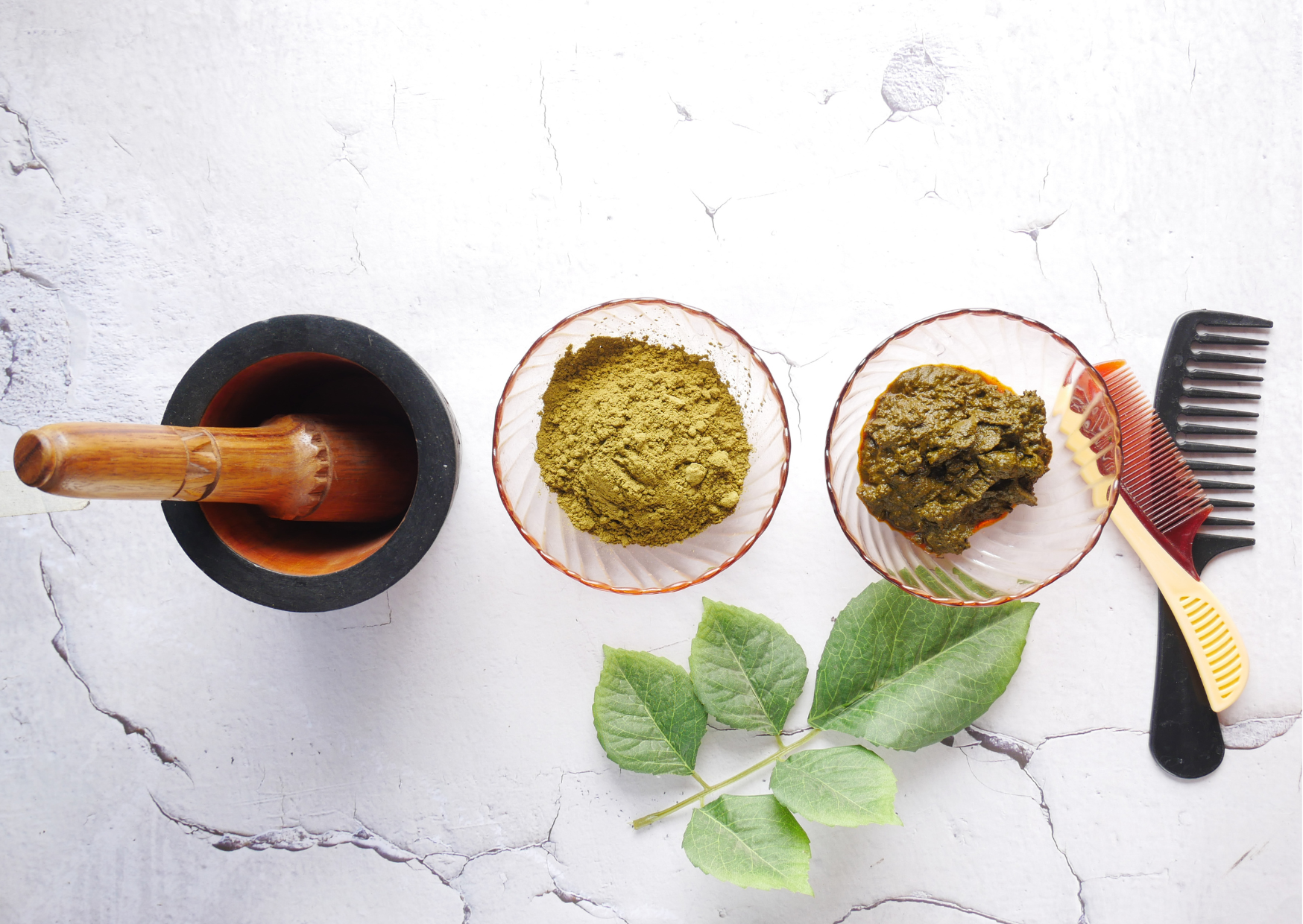a flat lay lifestyle image of it's pure organic herbal hair dyes. from left to right you can see a wooden and stone pestle and mortar, 2 glass bowls with a pink tint, one showing the dry powder which is an earthy green colour and the other showing the powder mixed with water to form a darker sludgy paste. there is one wooden and one plastic comb to the far right and a green leaf frond at the front of the row