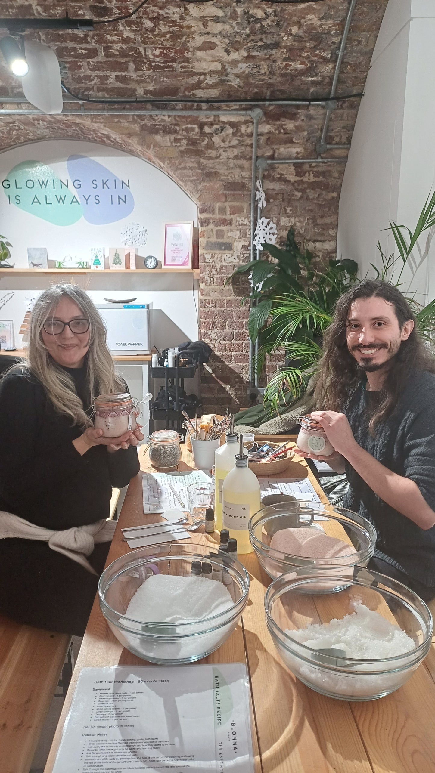 man and woman showing their finished bath salt creations after a workshop. on the table are the different kind of salts, carrier oils and essential oils that were used to make the bath salts