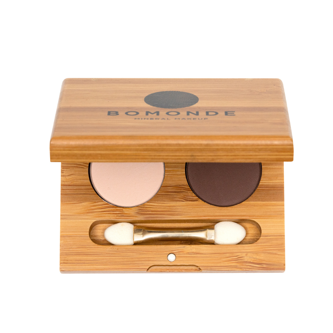 Aerial shot of micaline eyeshadow duo (nude and dark brown shadows). you can see the black text on the top of the bamboo refillable make up compact which reads 'bomonde mineral make up' with a partial view of the eyeshadows and brush