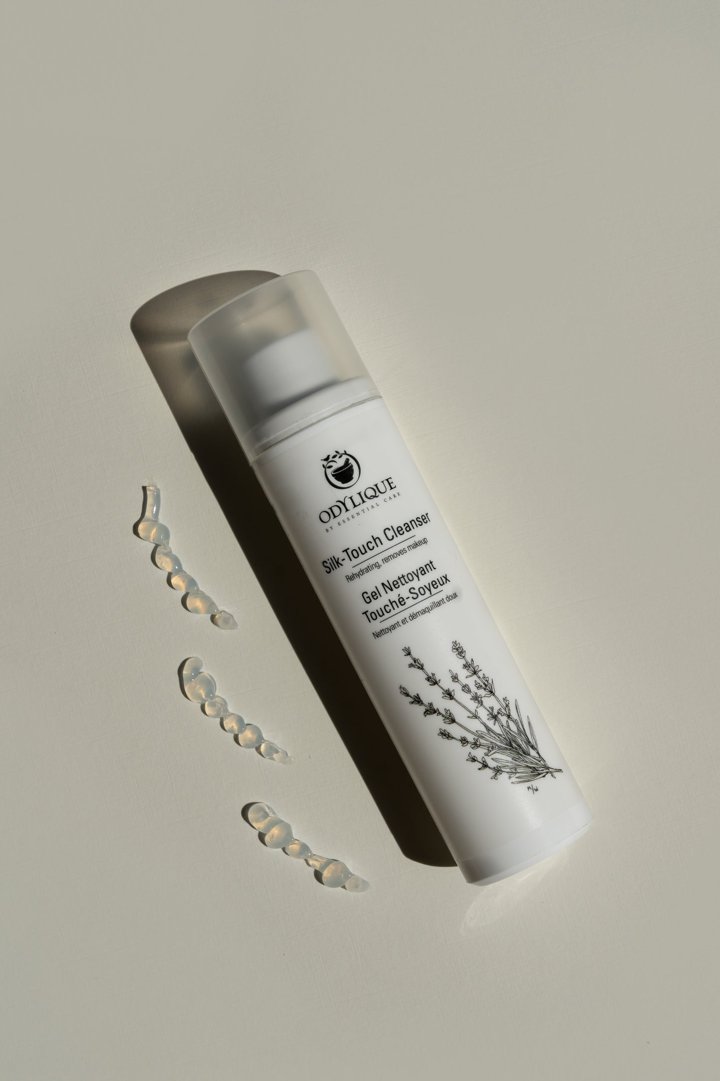 aerial view of odylique's silk touch cleanser on a light grey background. you can see the white pump bottle of the cleanser next to 3 lines of the gel cleanser dispensed on the surface. you can see the thick gel texture of the natural cleanser