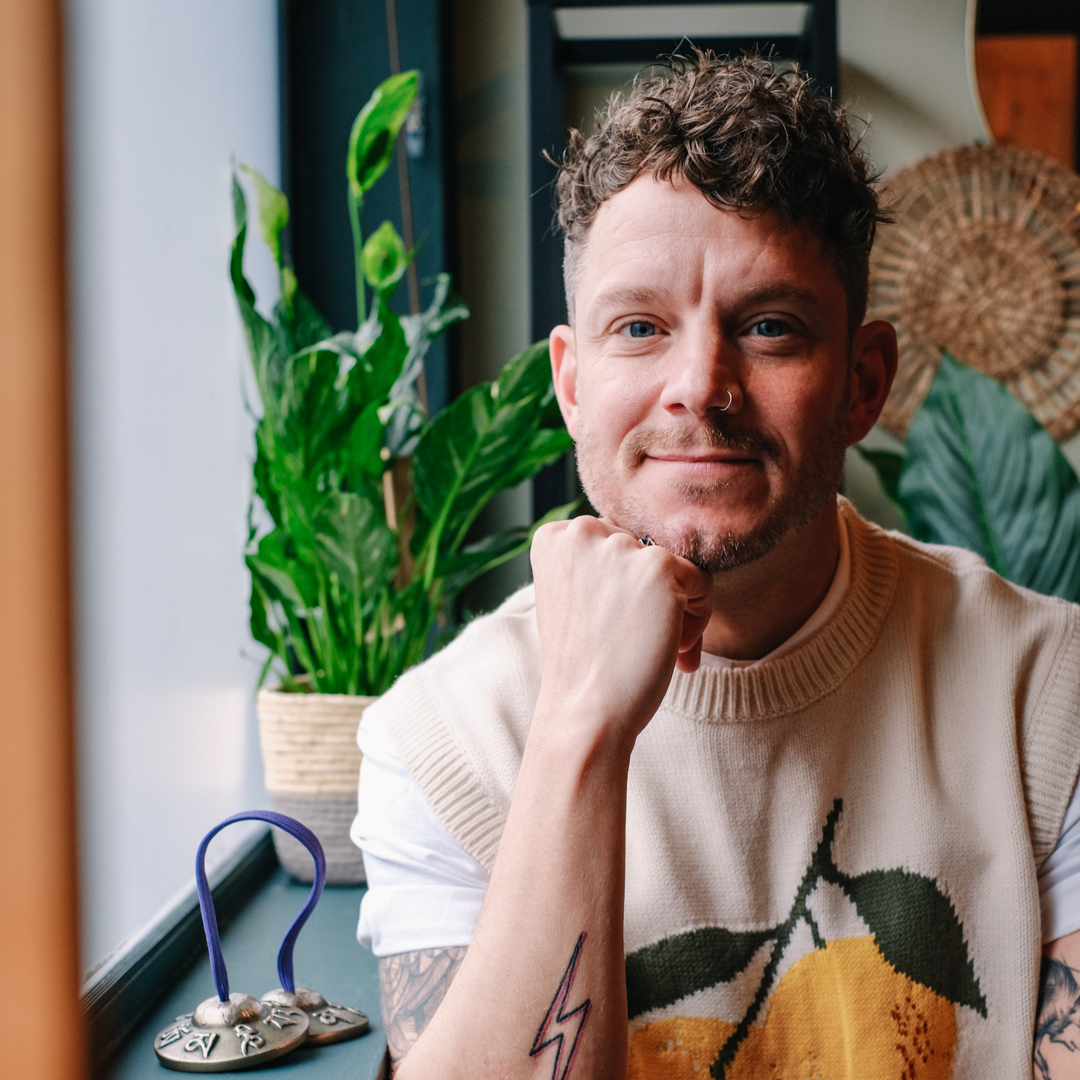ricky is sitting in his studio in stoke newington surrounded by geen plants, gongs and wicker wall hangings. he is wearing a white t shirt and a tank top with a knitted lemon motif