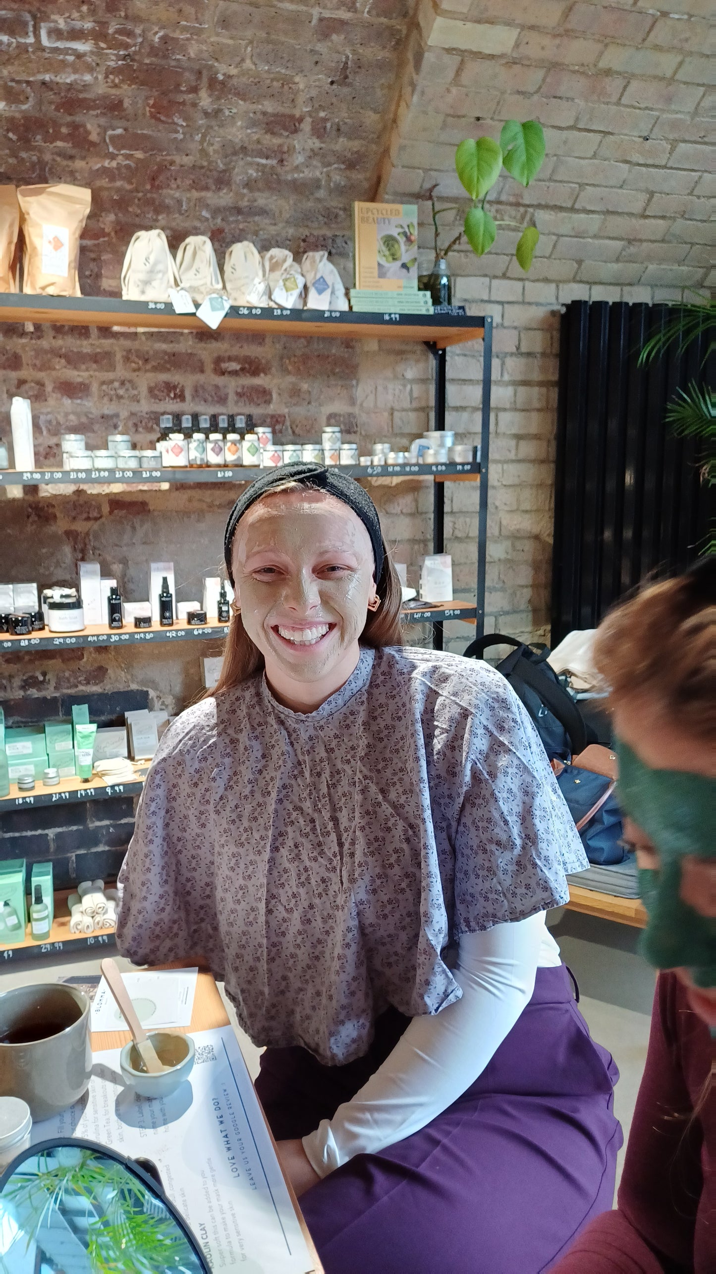 woman with fair hair wearing a black head band and floral cape smiling as she has applied her clay face mask in our natural skincare workshop in london